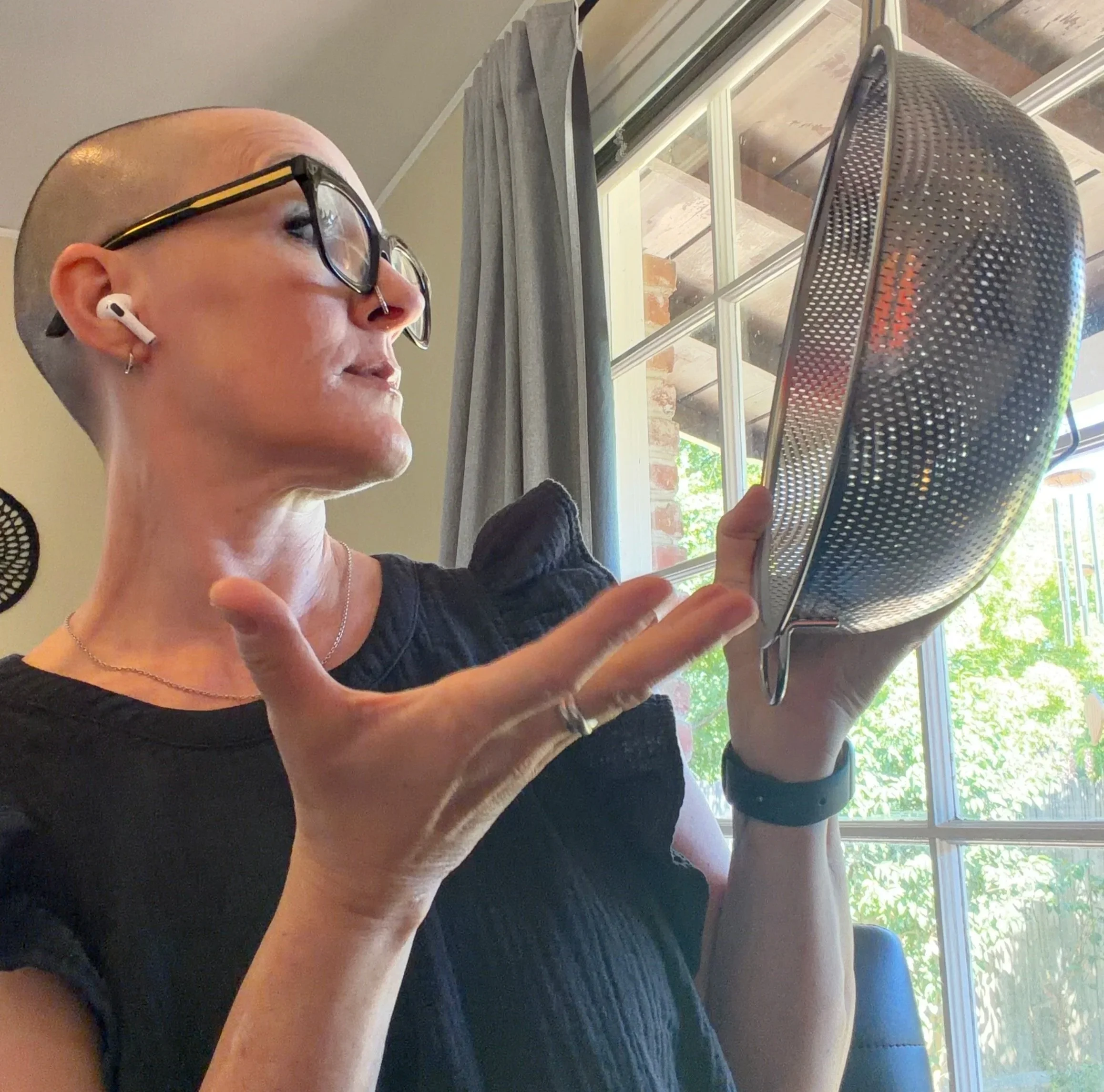 A woman with glasses and earbuds sitting near a window, reading or holding a device, with a view of a porch and trees outside.