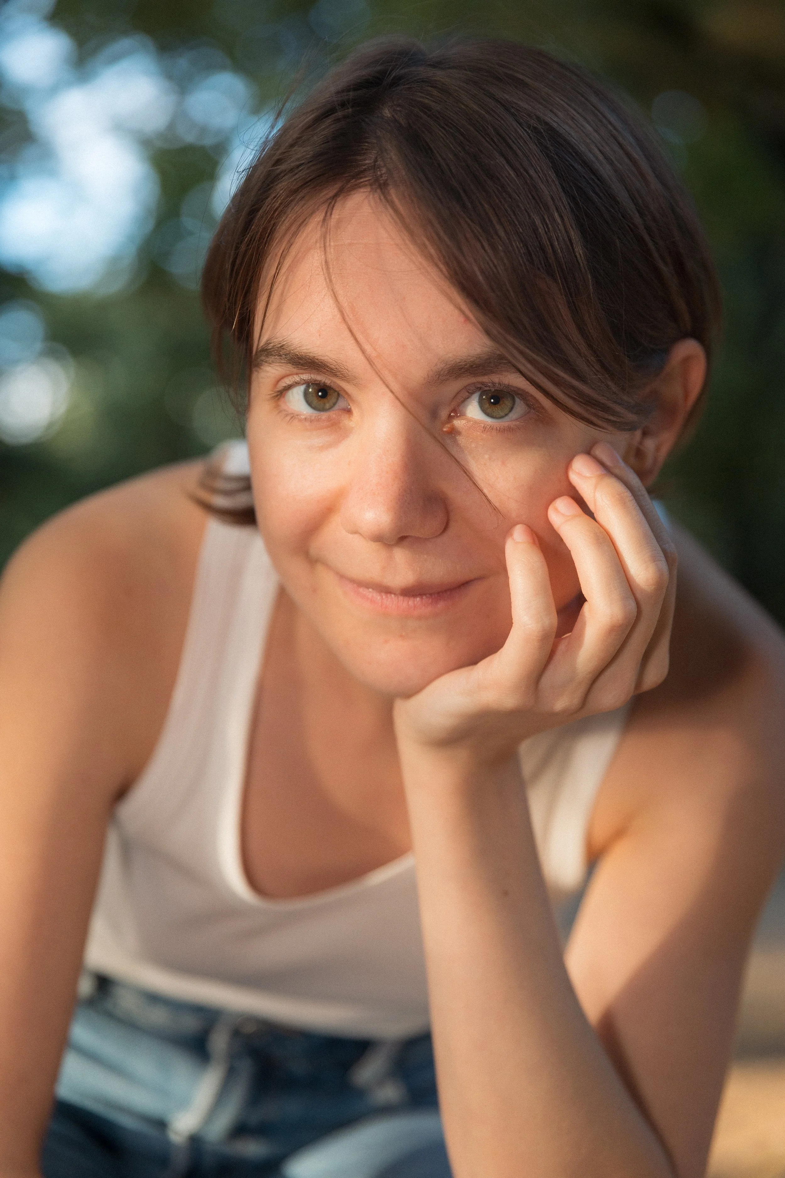 Photo Modelling Shoot in the Jardin de Luxembourg in Paris. Photo captured by Owen Clarke. Close-up of a woman with short brown hair, wearing a white sleeveless top, resting her chin on her hand, outdoors with green bokeh background.