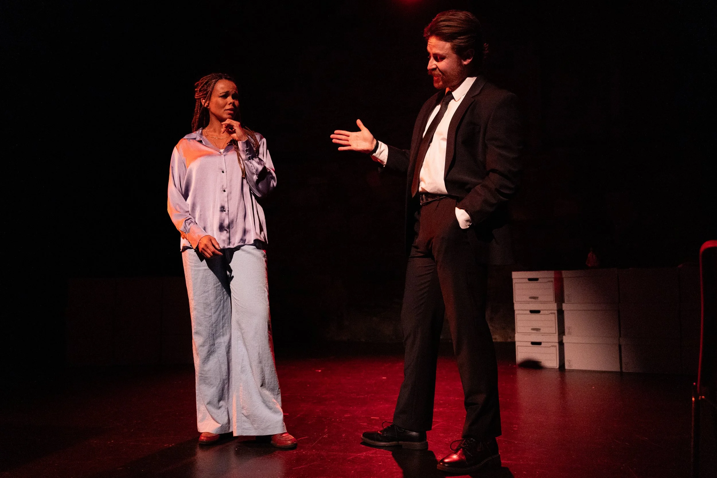 Two actors performing a scene on stage, with one woman in a satin blouse and wide-leg pants looking concerned, and a man in a suit gesturing with his hand.