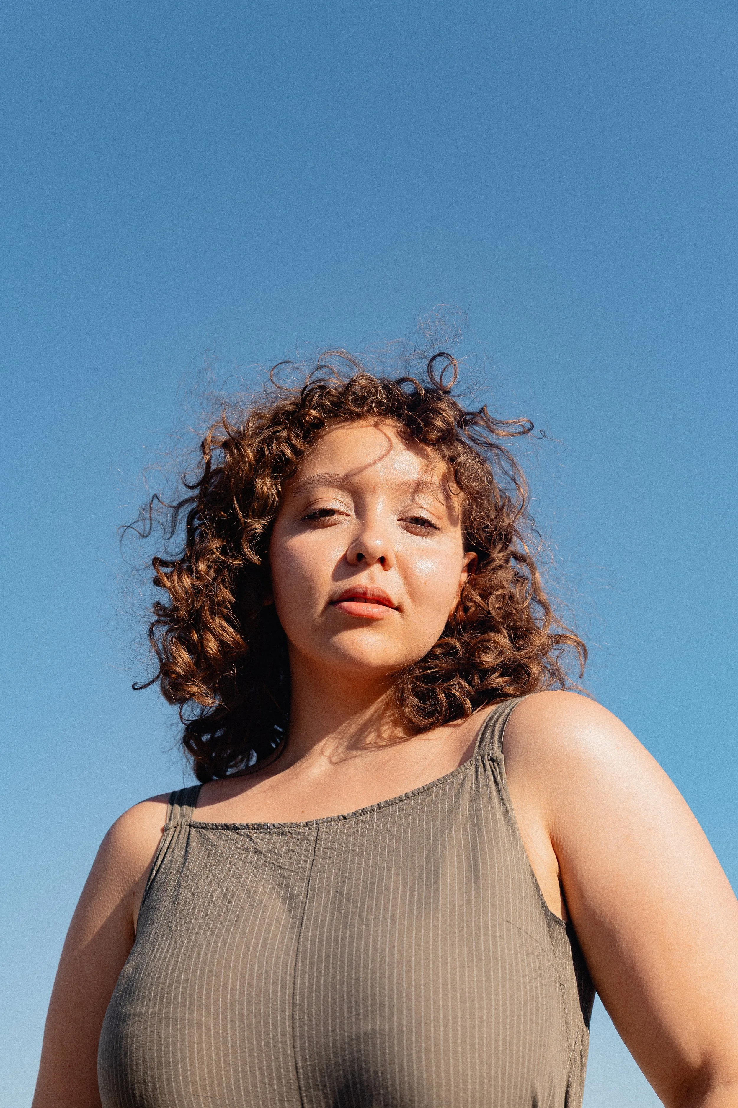 Photo Modelling Shoot in Paris during summertime. Photography by Owen Clarke.  A woman with curly hair standing outdoors against a clear blue sky, wearing a sleeveless top.