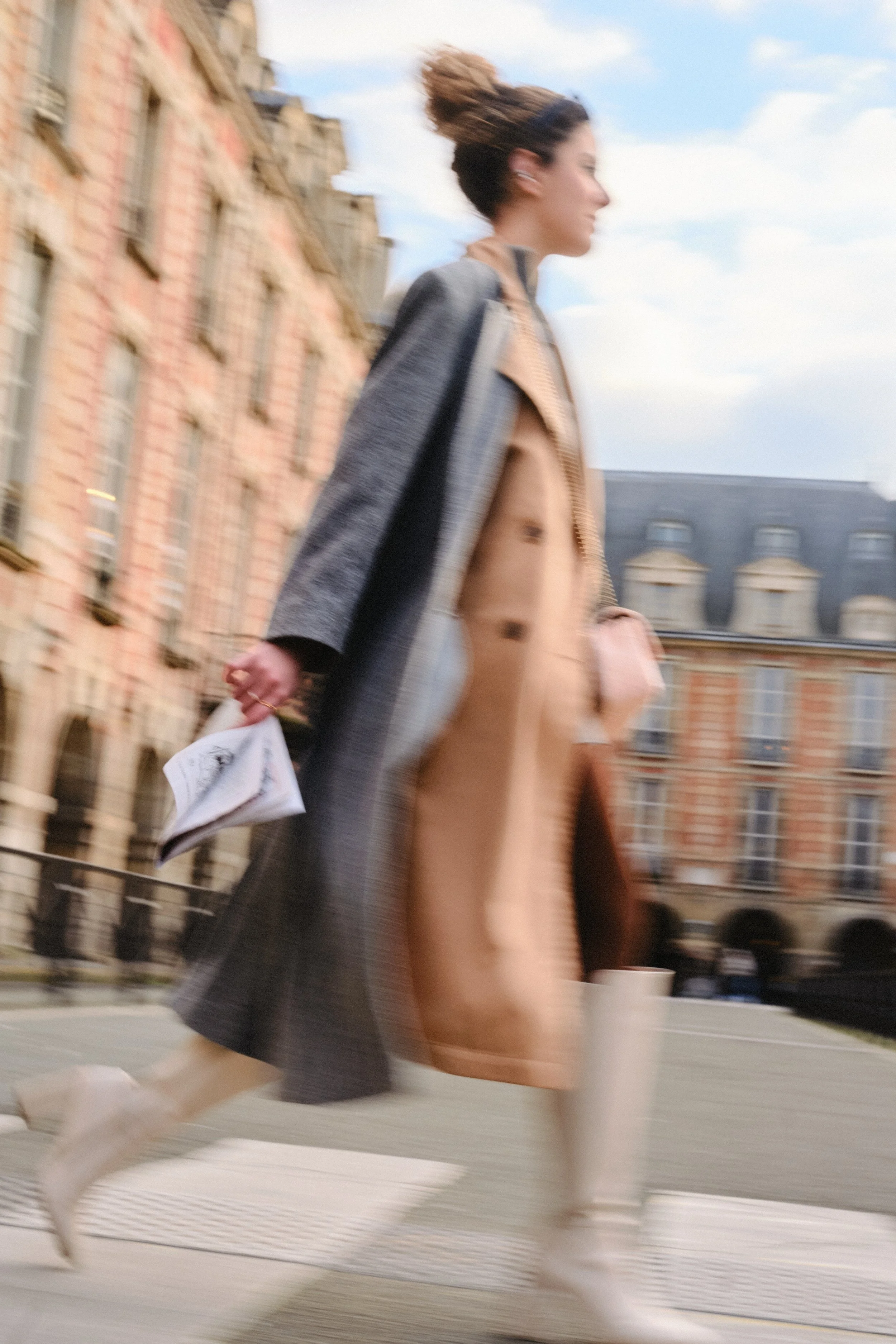 Photo of woman walking in Marais, Paris. Photography by Owen Clarke. Woman is wearing sunglasses, long grey coat, resting on a stone structure