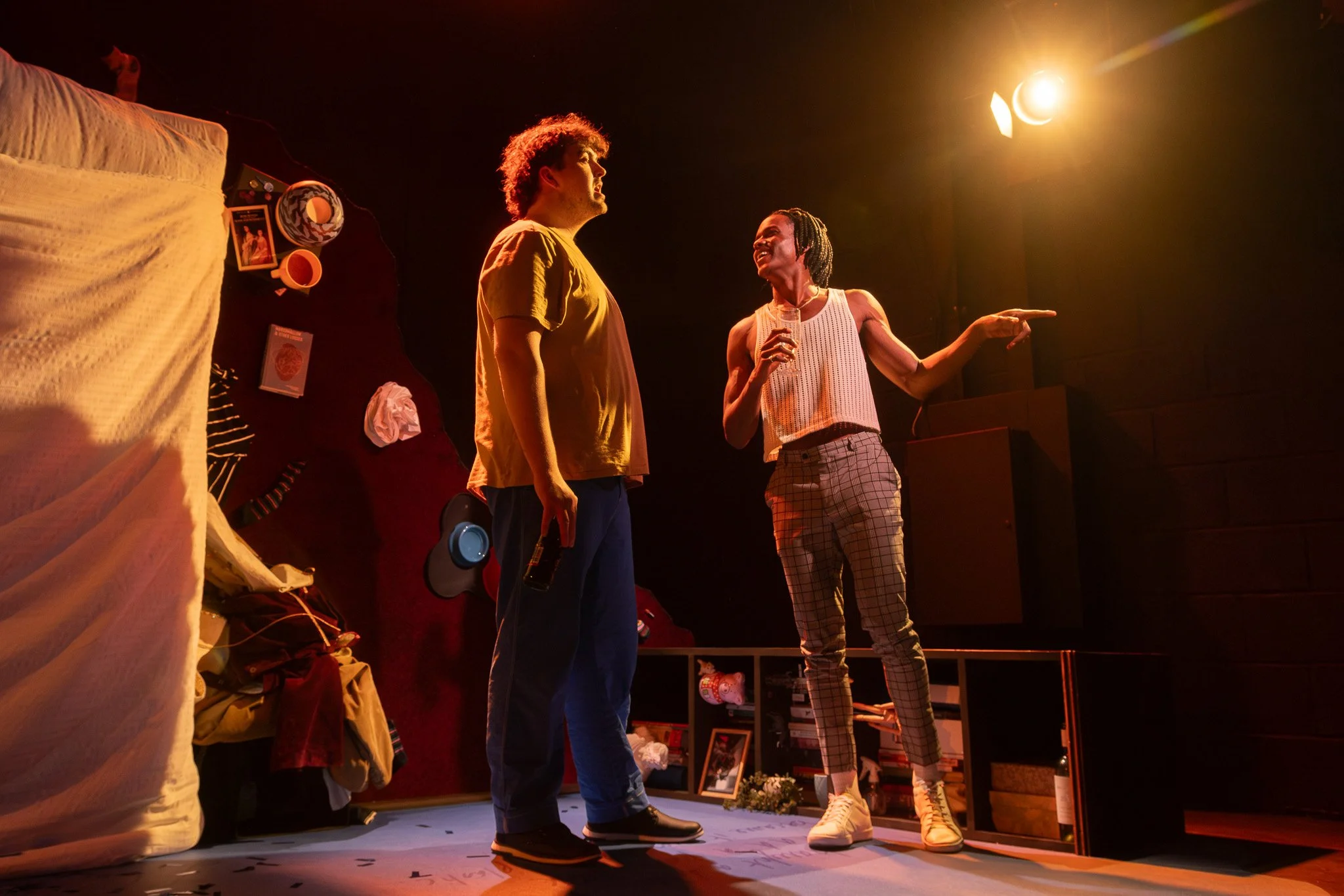 Two actors on stage during a performance, one man and one woman, with dramatic lighting and set decorations including a bed and various objects.