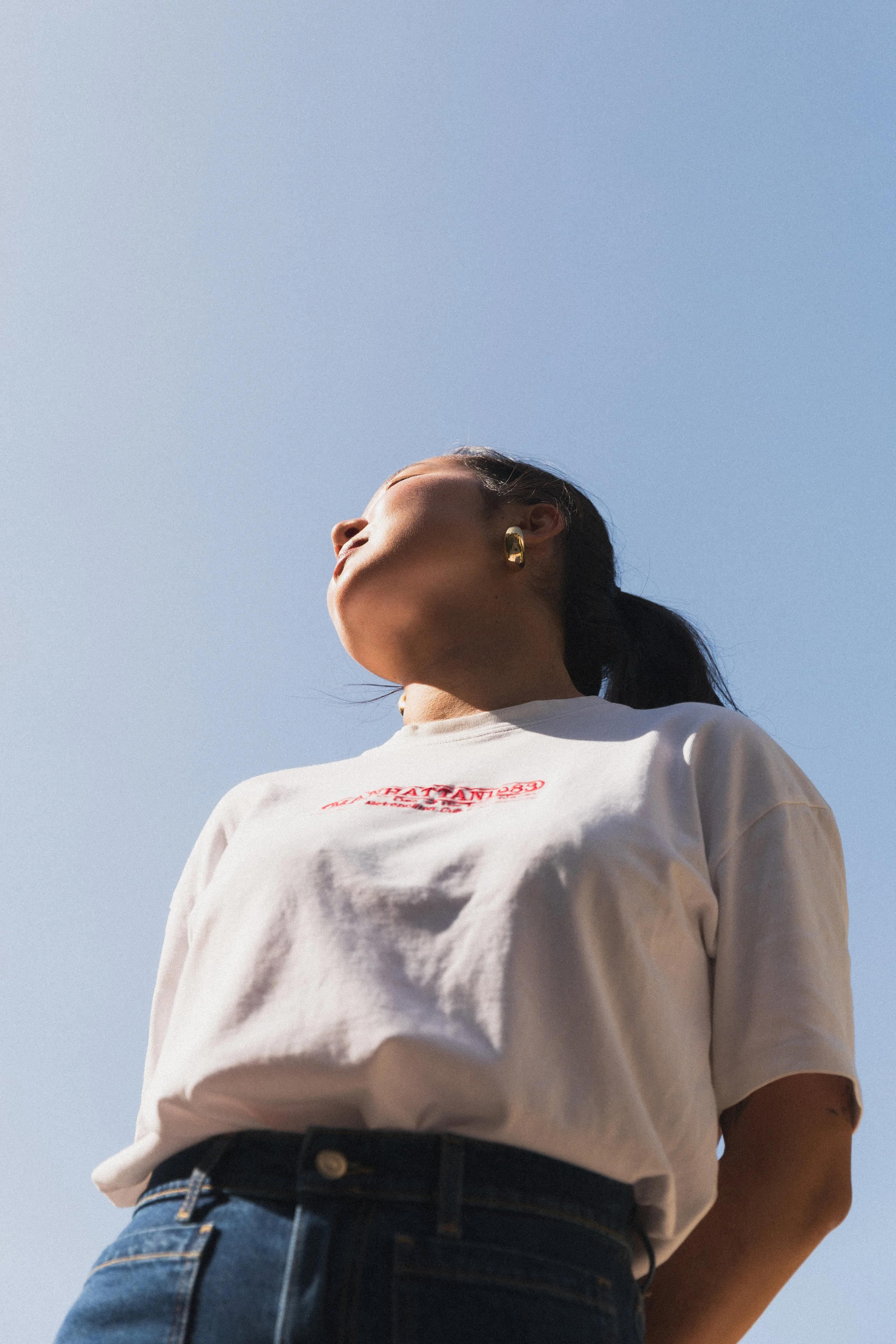 Photo Modelling Shoot in the Marais, Paris. Photography by Owen Clarke. Low-angle view of a woman with dark hair tied back, wearing a white t-shirt with red text and dark jeans, standing outdoors against a clear blue sky.