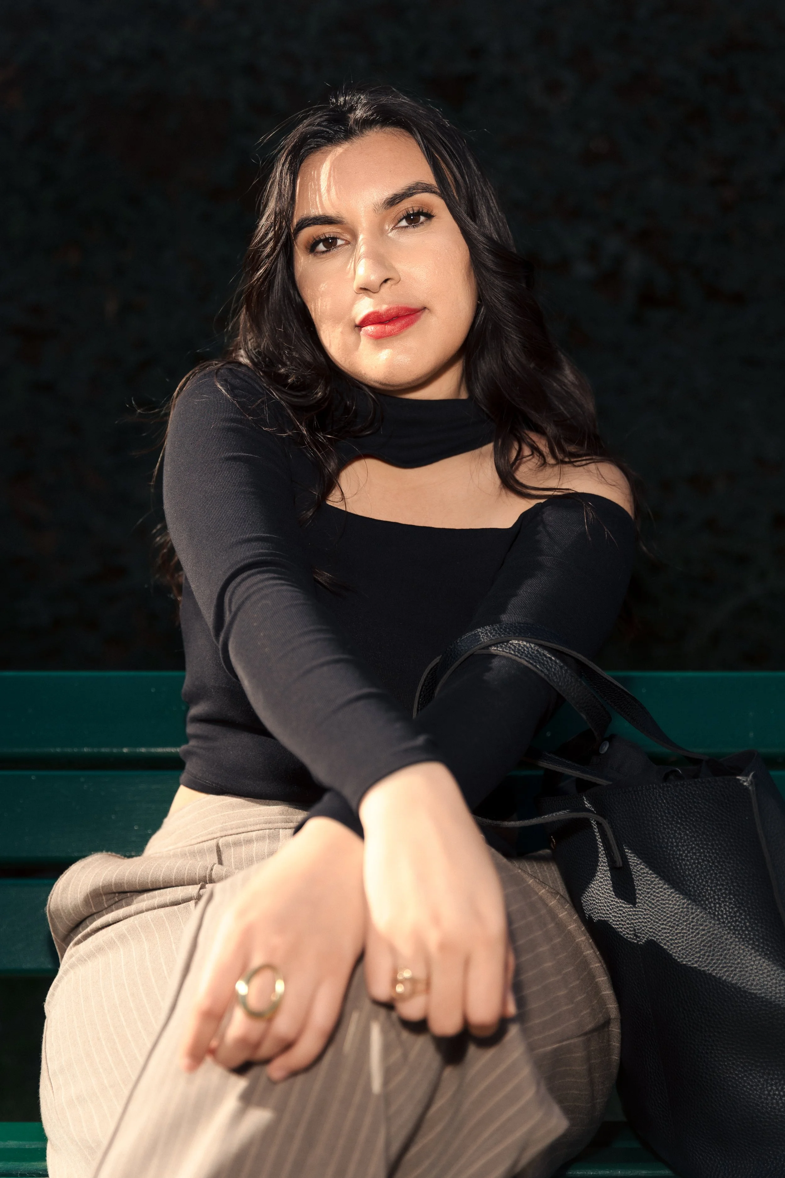 Photoshoot in Paris. Photography by Owen Clarke. Woman is wearing a black top, beige trousers, sitting in on a green bench infront of a green hedge