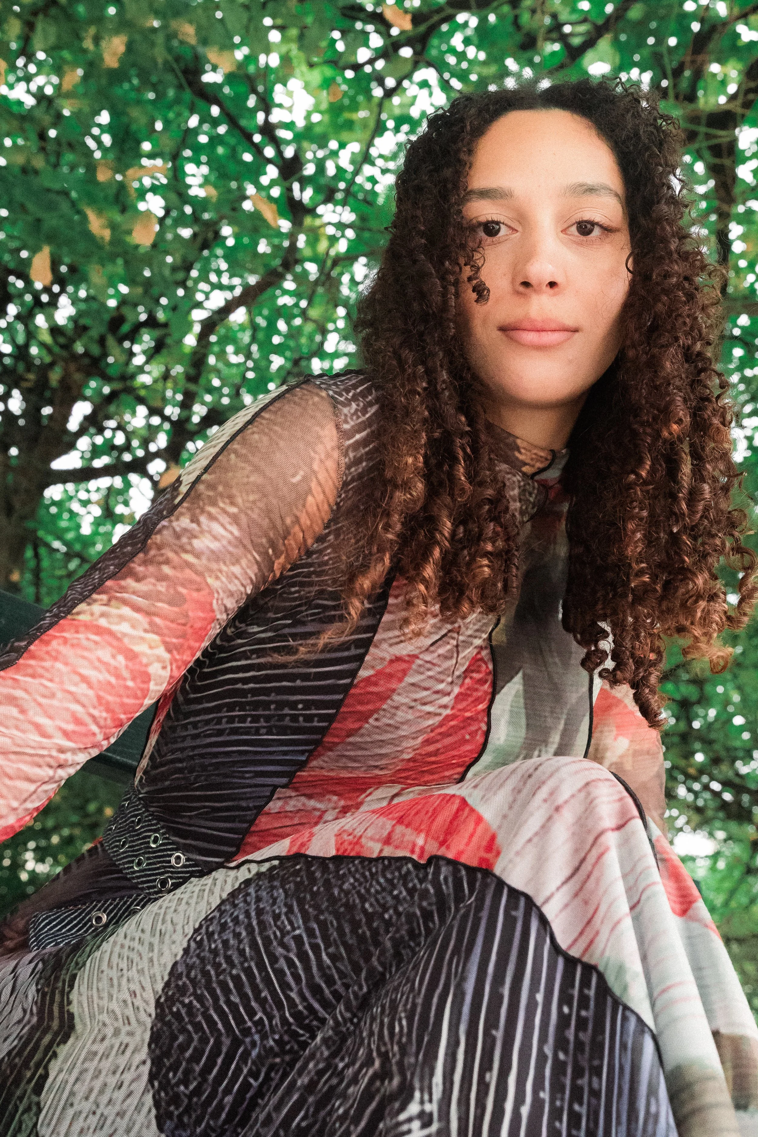 Photo Modelling Shoot in the Marais, Paris during summertime. Photography by Owen Clarke. The woman with curly brown hair sitting outdoors beneath a leafy tree, wearing a multicolored patterned dress.