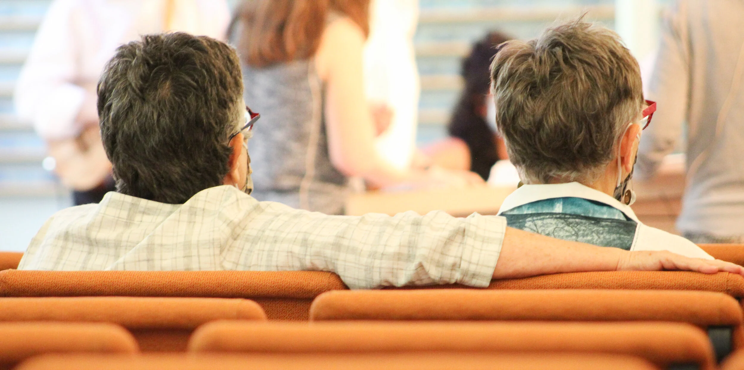Two older men with gray hair and glasses sitting on an orange bench, one has his arm around the other. They are in a crowded indoor setting with other people in the background.