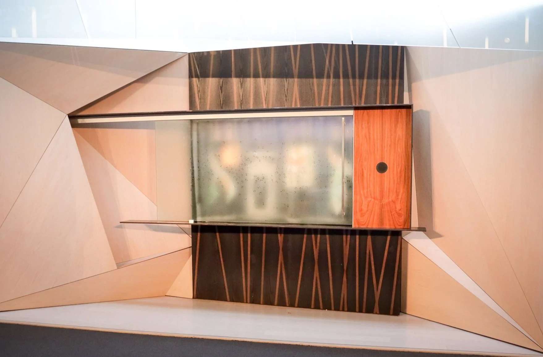 An abstract geometric wall installation with various wooden and metallic panels arranged in sharp angles, featuring a sliding door with a circular handle.
