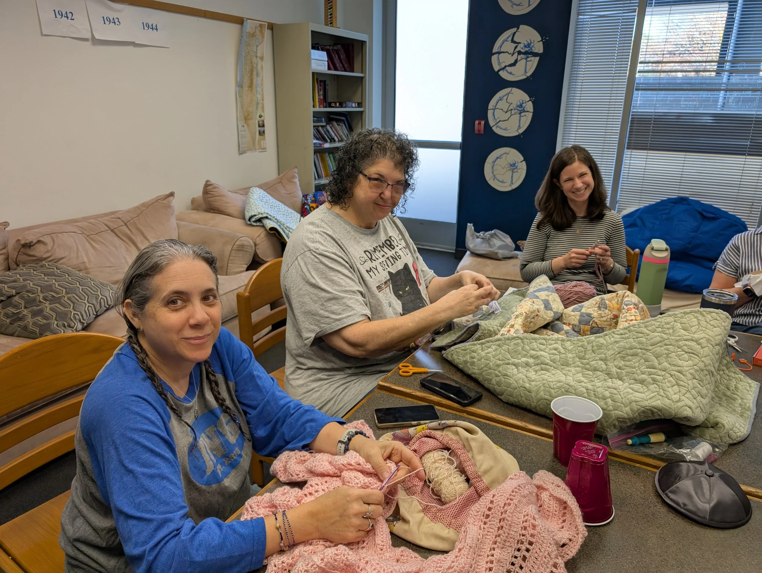 Four women sitting at a table working on knitting projects, with yarn and knitting tools in front of them, in a room with bookshelves and decorative plates on the wall.