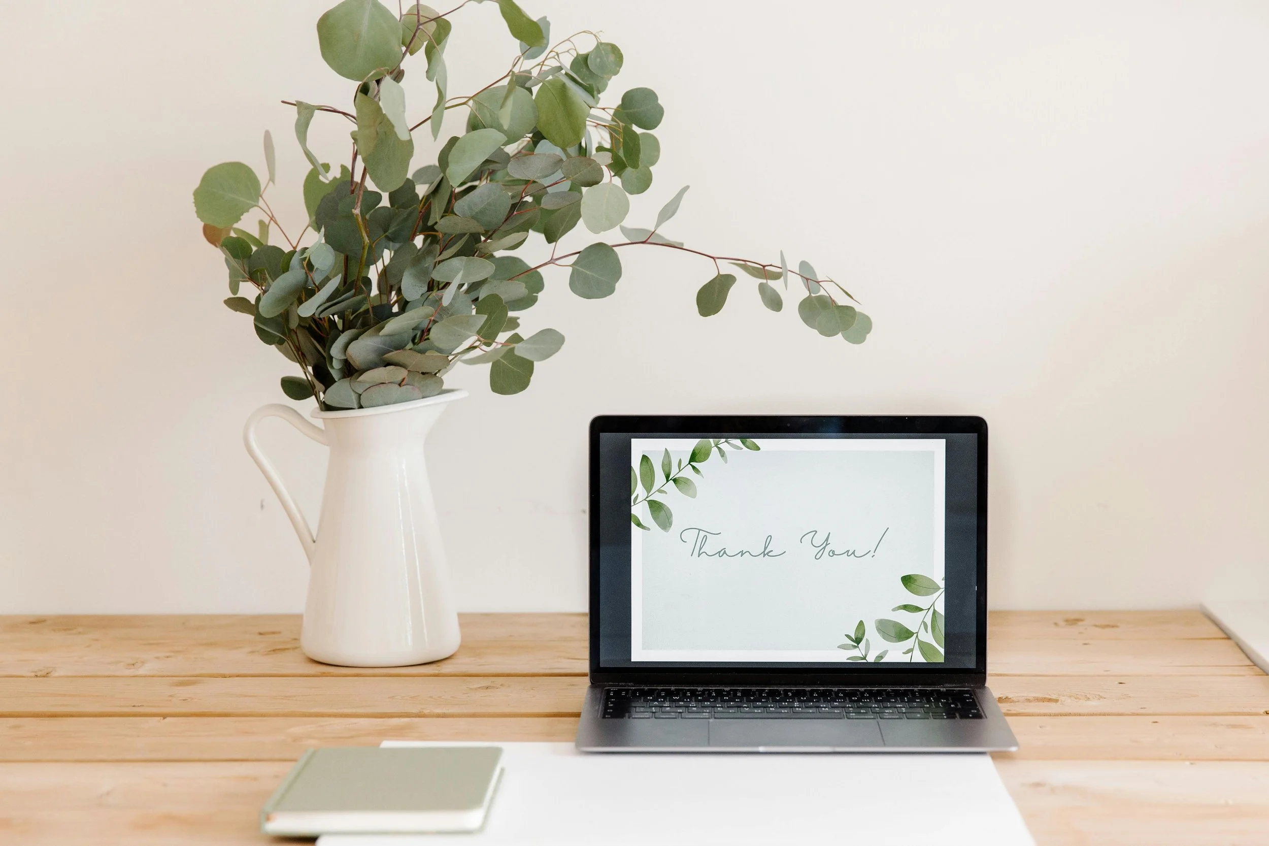 A desk with a laptop displaying a 'Thank You!' message, a closed notebook, and a large vase with green eucalyptus branches.