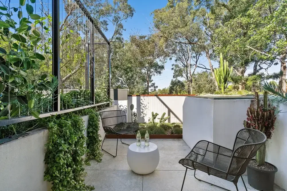 A cozy outdoor patio with two black metal chairs, a small white round table with two glass bottles, surrounded by potted and hanging green plants, and lush trees in the background under a clear blue sky.