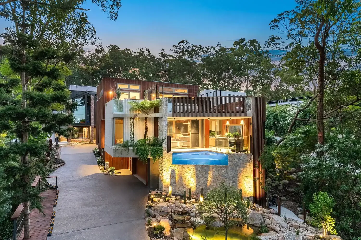 Modern multi-story house with wood and stone exterior, large glass windows, and outdoor pool at dusk, surrounded by lush greenery and trees.
