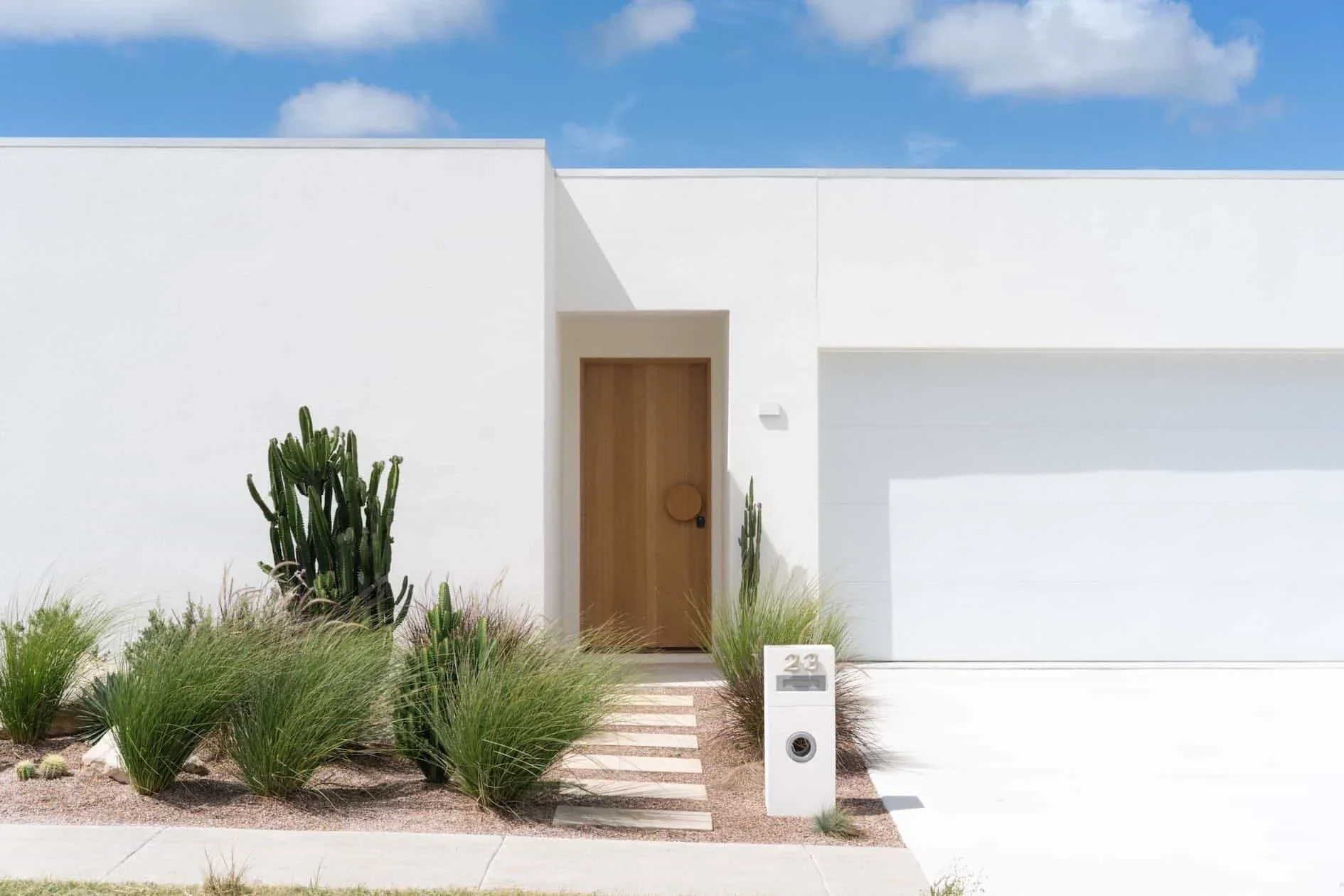 Minimalist white modern house with a wooden front door, desert landscaping including cacti and grasses, blue sky with some clouds.