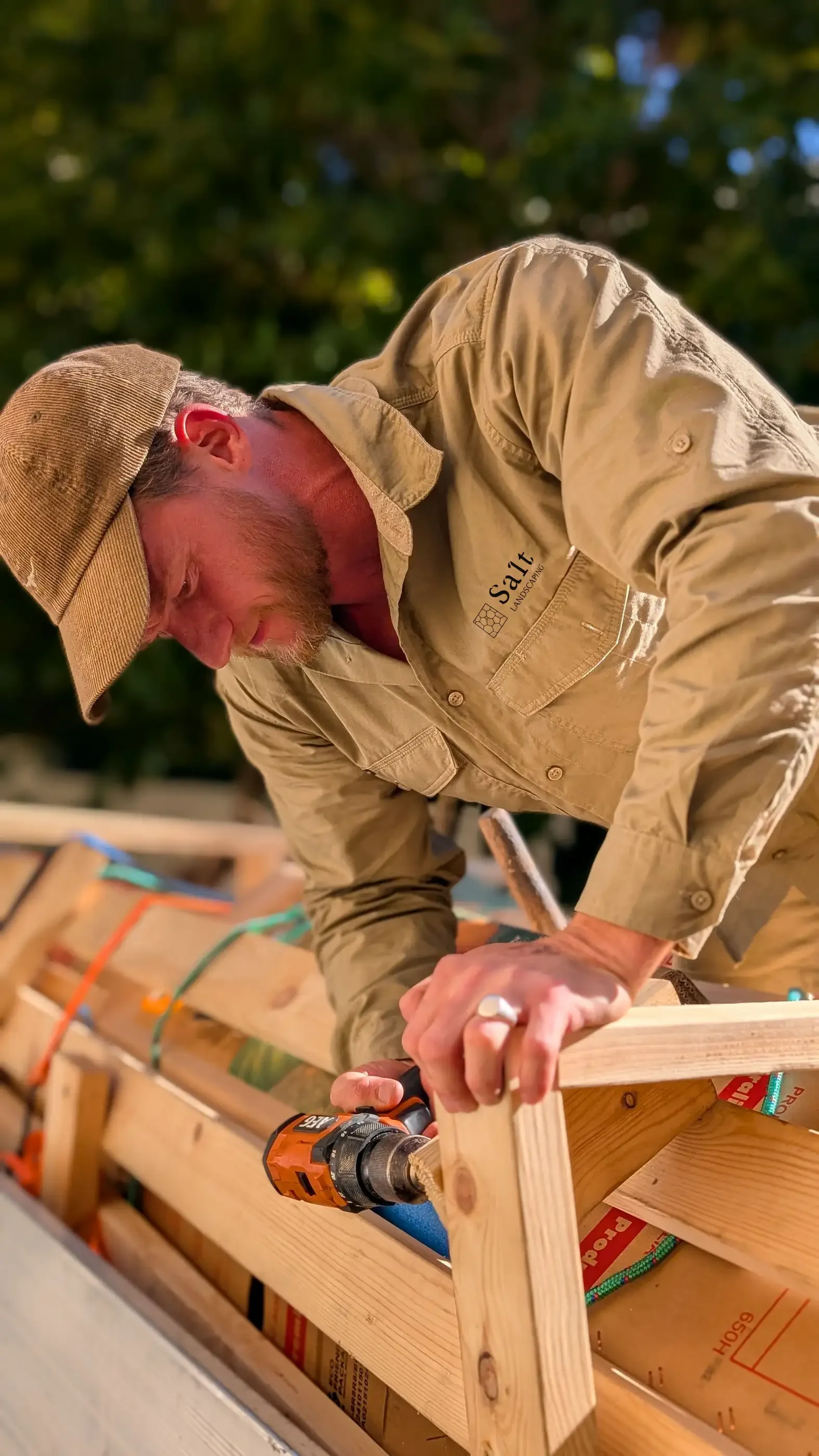 A man wearing a beige work uniform and a wide-brimmed hat is assembling a wooden structure outdoors using a cordless drill.