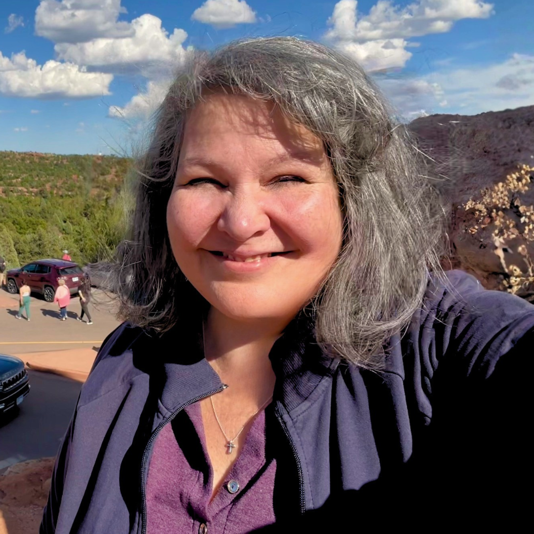 Portrait of a woman with gray hair, wearing a black and white patterned top and green cardigan, smiling outdoors.
