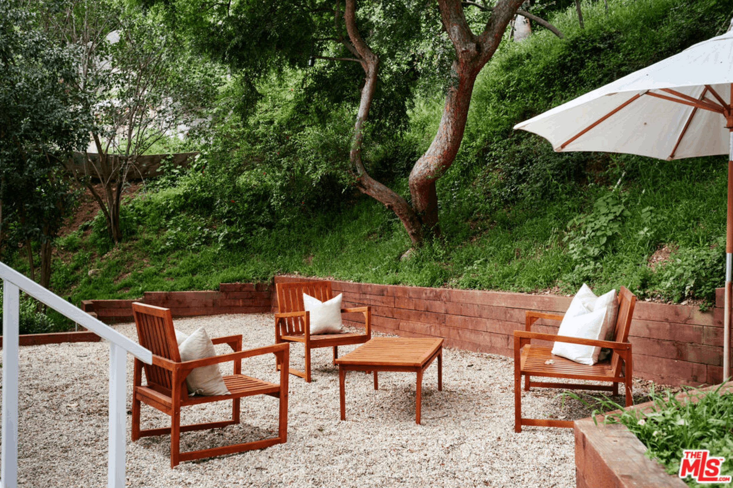 A backyard patio area with wooden chairs and pillows, a white umbrella, and a lush green hillside with trees and bushes.