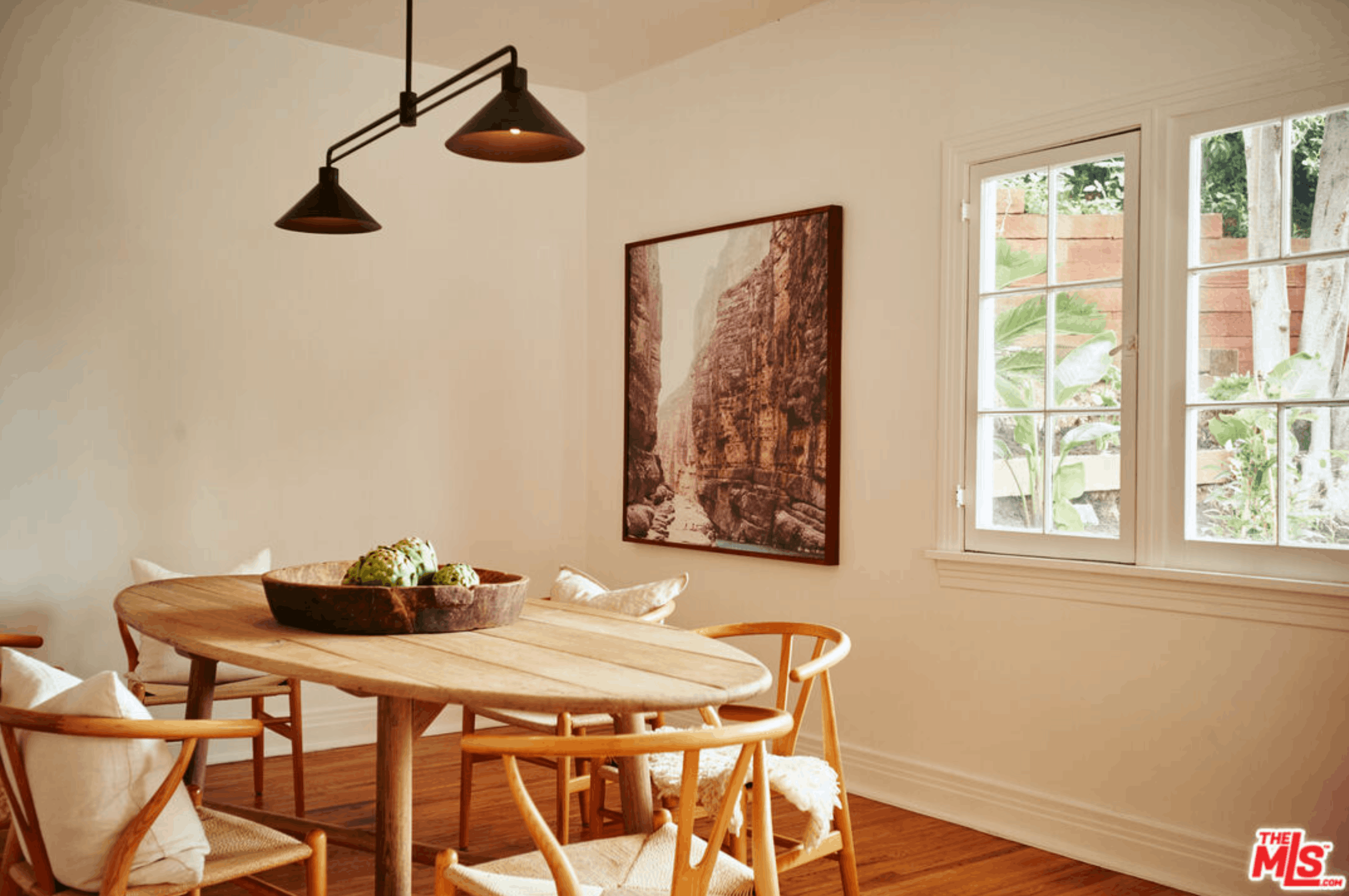 A dining area with a wooden round table, surrounded by matching wooden chairs with white cushions. The table holds a dark bowl filled with green decorative objects. There is a large rectangular wall art depicting a canyon or rocky landscape hanging on the beige wall. Natural light streams in through a paneled window revealing trees and a wooden fence outside. Two black cone-shaped pendant lights hang above the table.