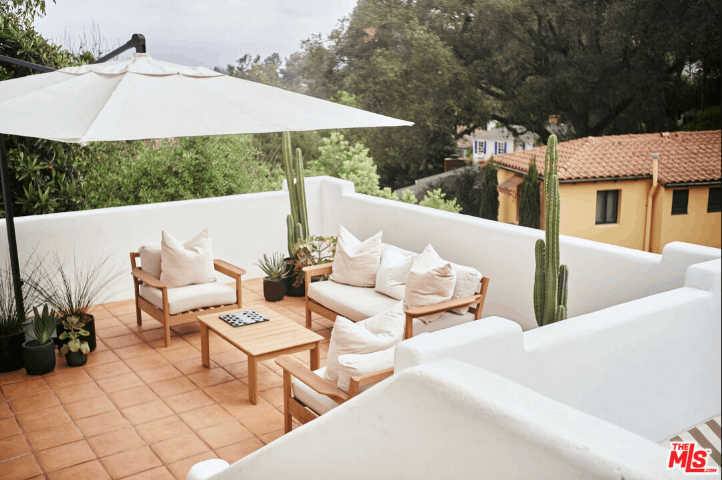 An outdoor patio with white cushioned furniture, including chairs and a sofa, wooden tables, various potted plants, and tall cacti. A white patio umbrella provides shade, and there are trees and a building with a red tile roof in the background.