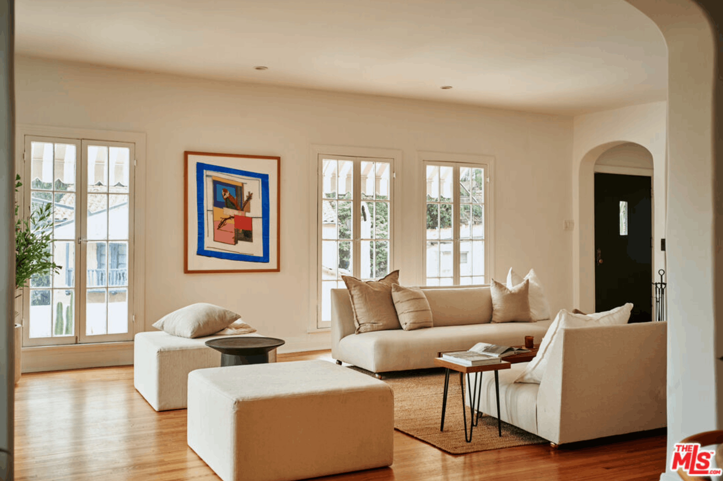 A living room with beige couches, a black side table, and framed artwork on the wall. Large windows let in natural light, with wooden flooring and a white wall. A doorway leads to another room.