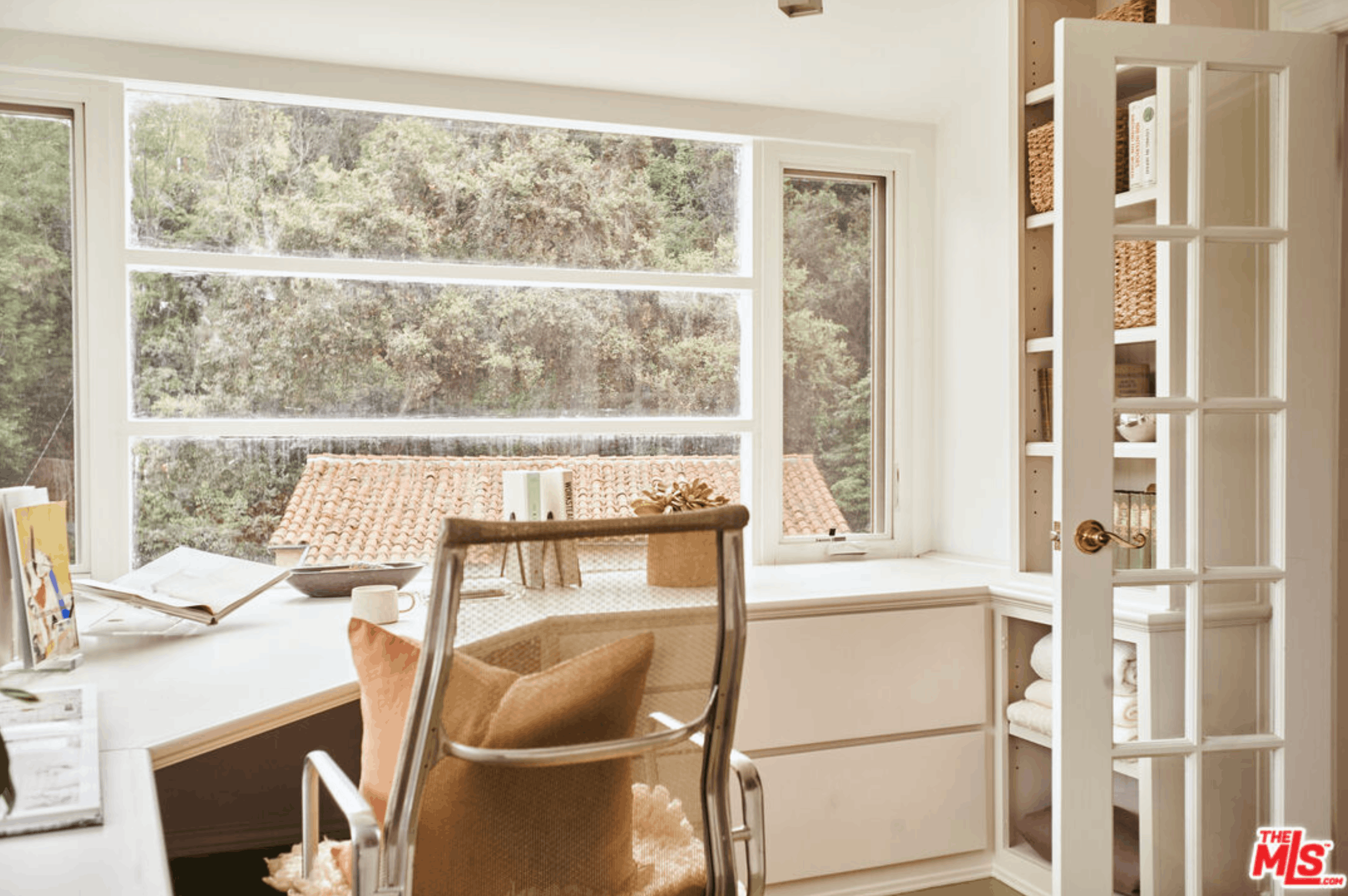 A cozy corner of a room with a large window showing trees outside. There is a white table with books, a bowl, and a cup, and a tan woven chair pushed in. To the right, a white bookshelf contains baskets and books, and a small decorative item is visible on top. The room is bright and inviting with natural light.
