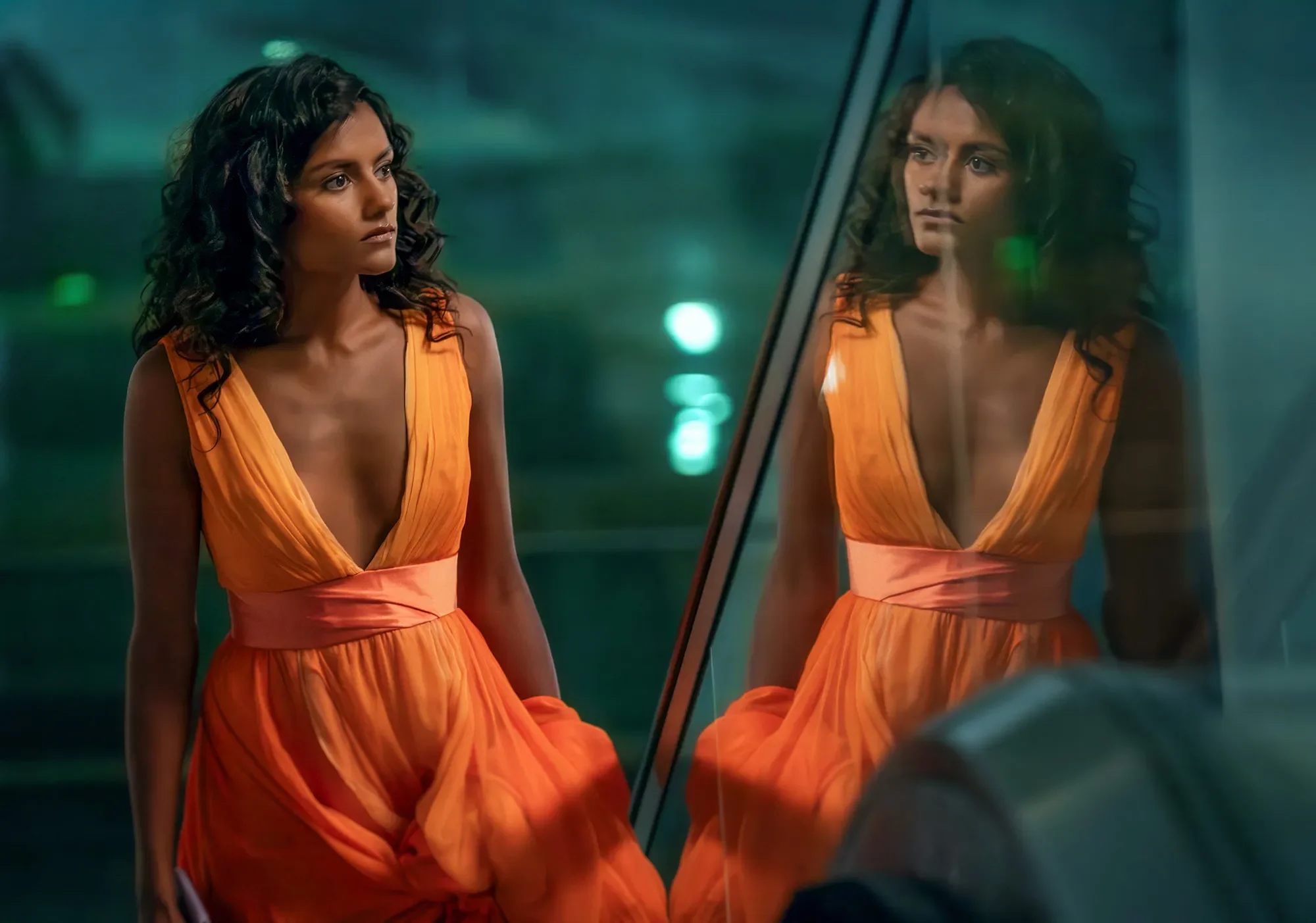 A woman with dark curly hair wearing a sleeveless orange dress looking at her reflection in a mirror.