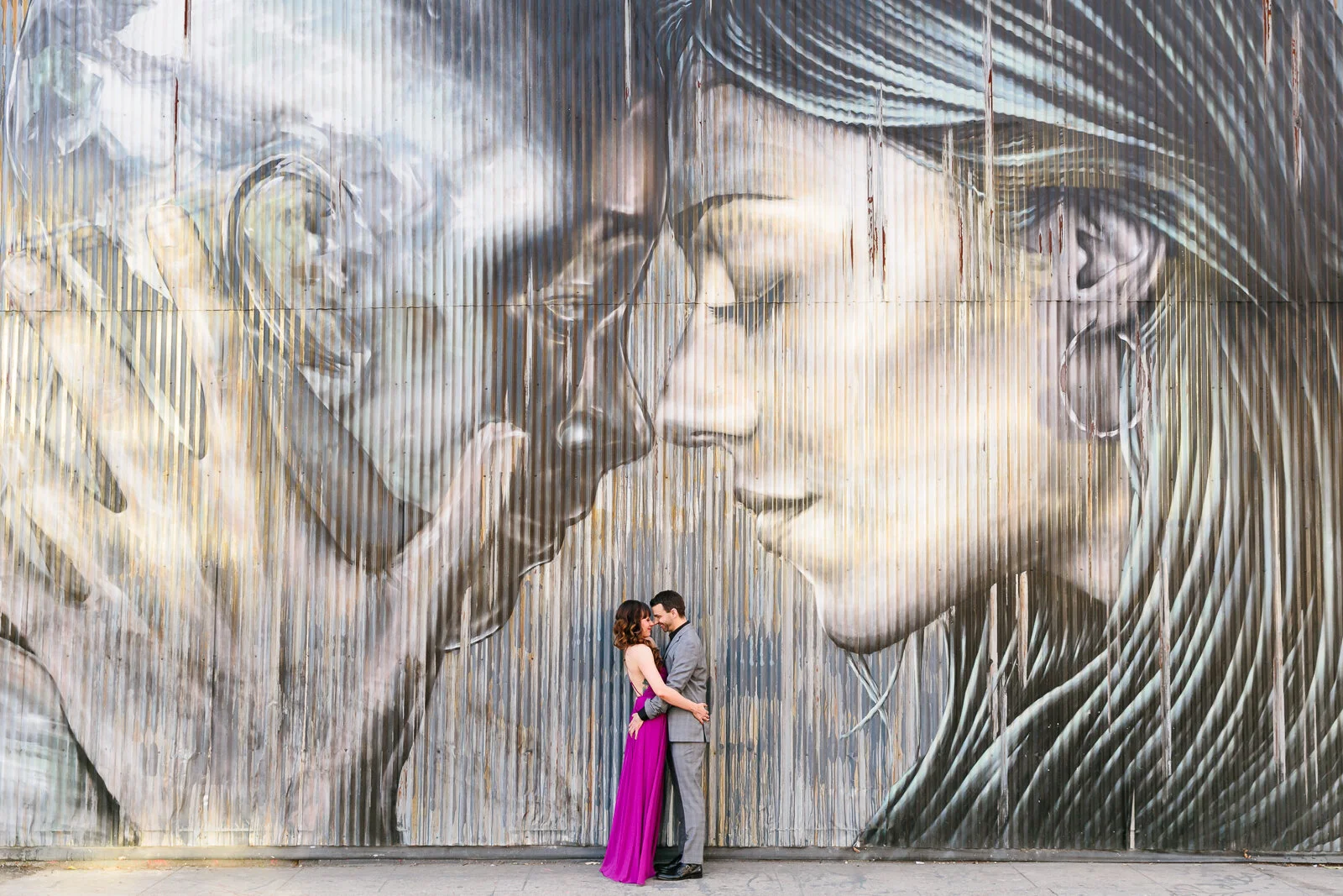 Couple embracing in front of a large mural of two faces on a corrugated metal wall.