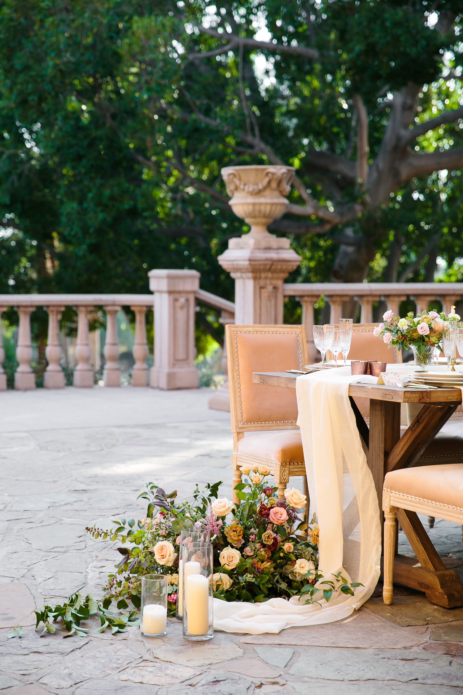 Outdoor patio with a wooden table and beige chairs, adorned with floral arrangements and candles. A draped white cloth cascades to the stone floor.