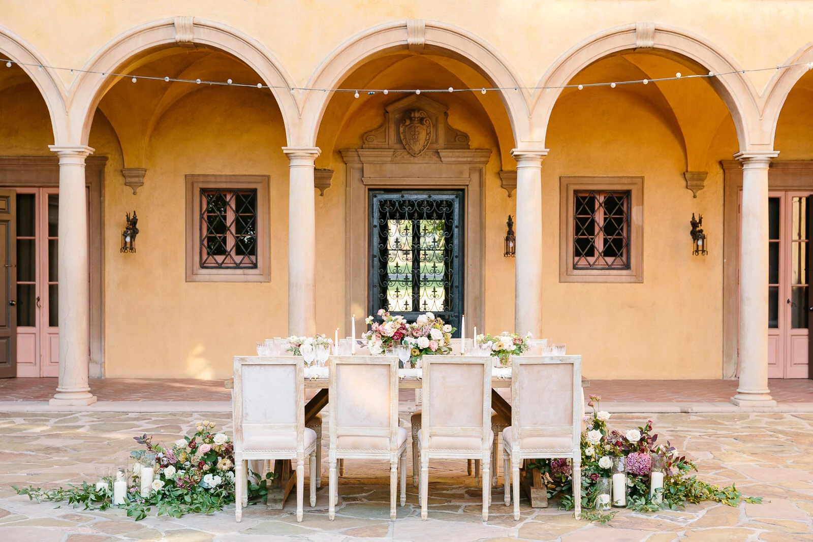 Elegant outdoor dining setup in a courtyard with arched columns, featuring a long table decorated with flowers and candles, surrounded by white chairs on a stone patio.