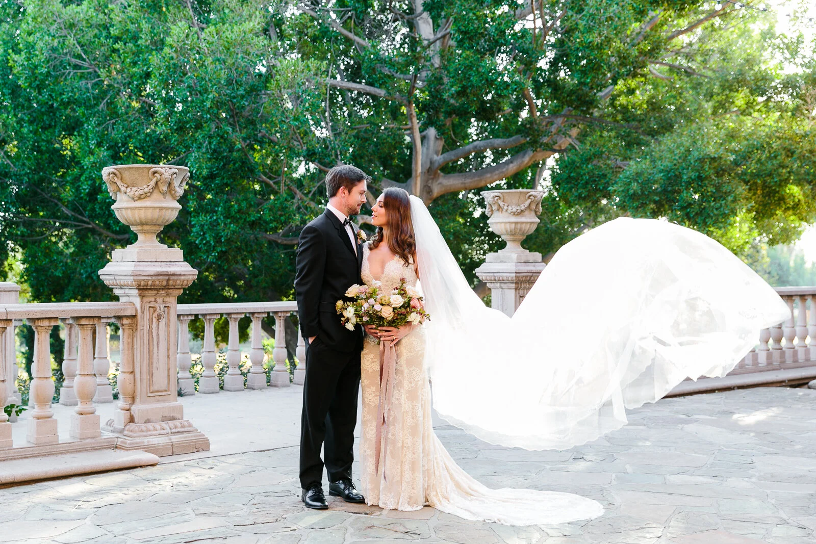 Bride and groom standing outdoors in formal attire, with the bride's veil flowing and greenery in the background.