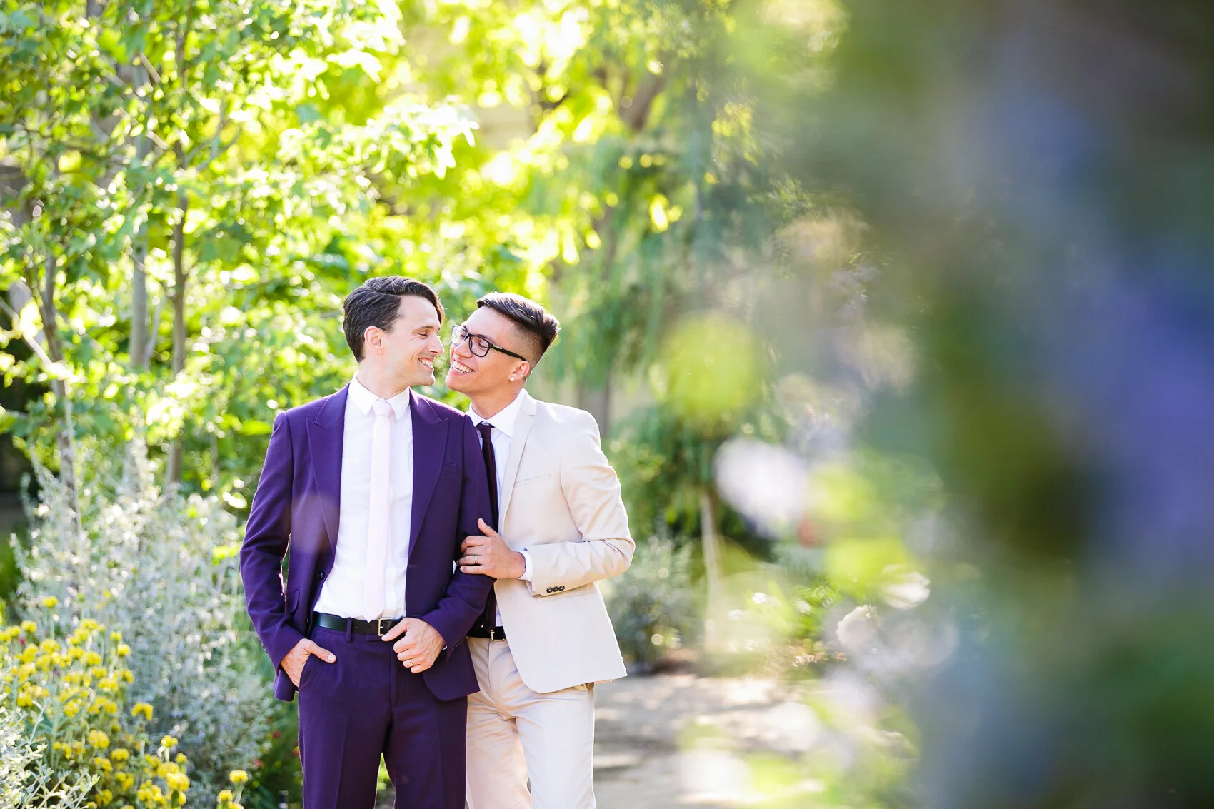 Two men in suits embracing and smiling at each other in a lush garden setting.