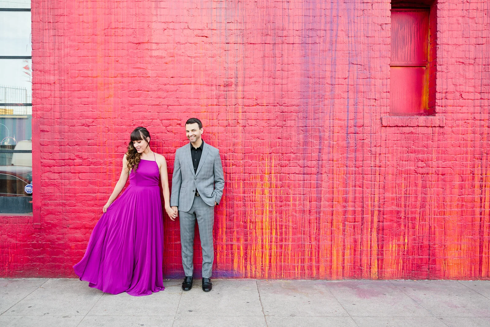 Couple holding hands in front of a red brick wall, with the woman in a purple dress and the man in a gray suit.