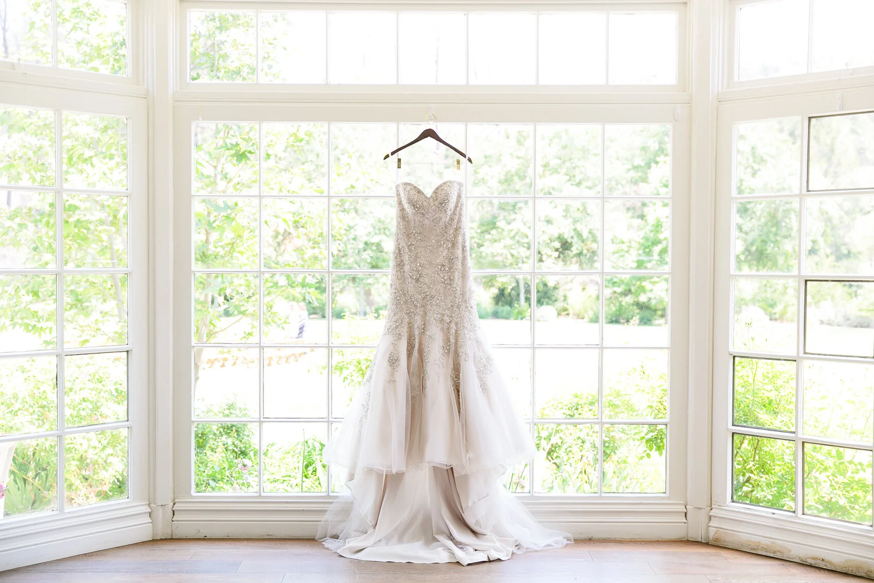 A wedding dress with intricate details hangs on a hanger in front of a large window, with a garden view in the background.