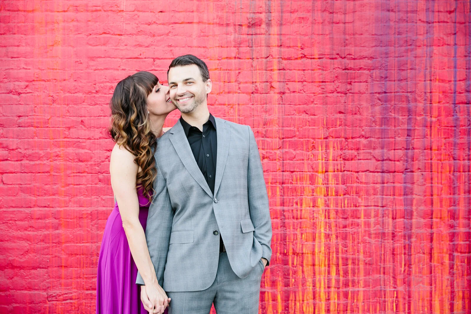 Couple posing in front of a red brick wall; woman in a purple dress kisses man in a gray suit.