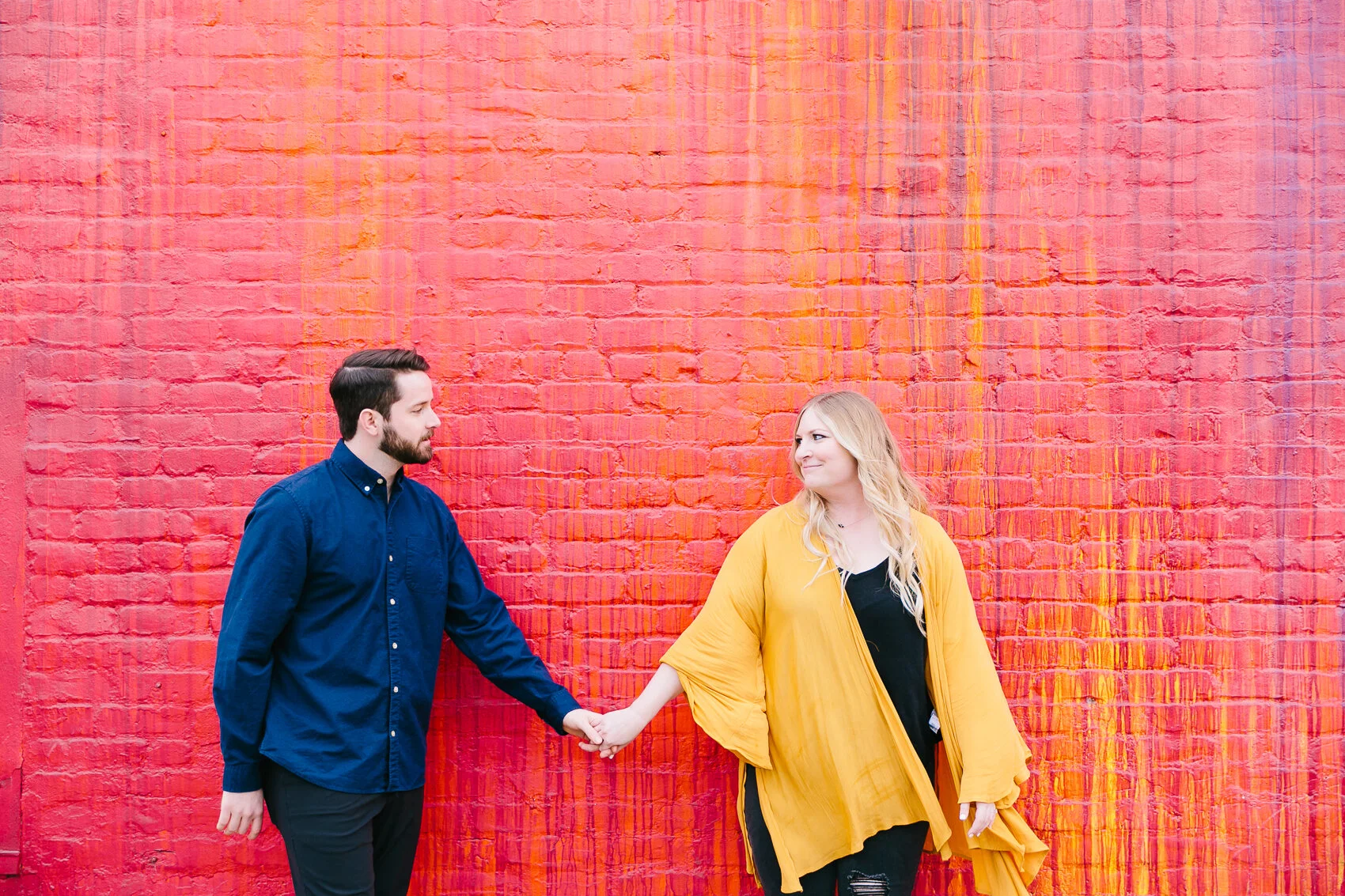 Couple holding hands in front of a vibrant red and orange brick wall.