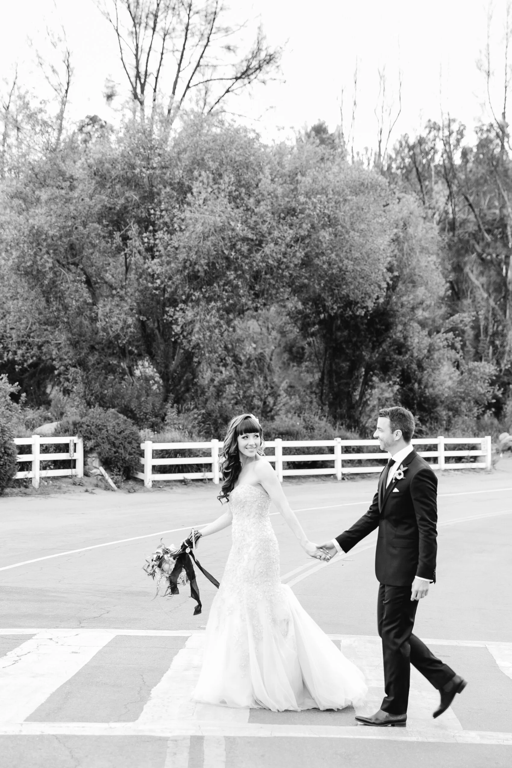 Black and white photo of a bride and groom walking hand in hand on a street, with trees and a white fence in the background. The bride is wearing a strapless gown and holding a bouquet, and the groom is in a suit.