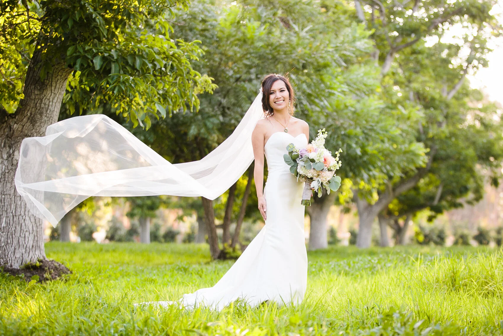 Bride in a white wedding dress holding a bouquet, standing in a green outdoor setting with trees, veil flowing in the wind.