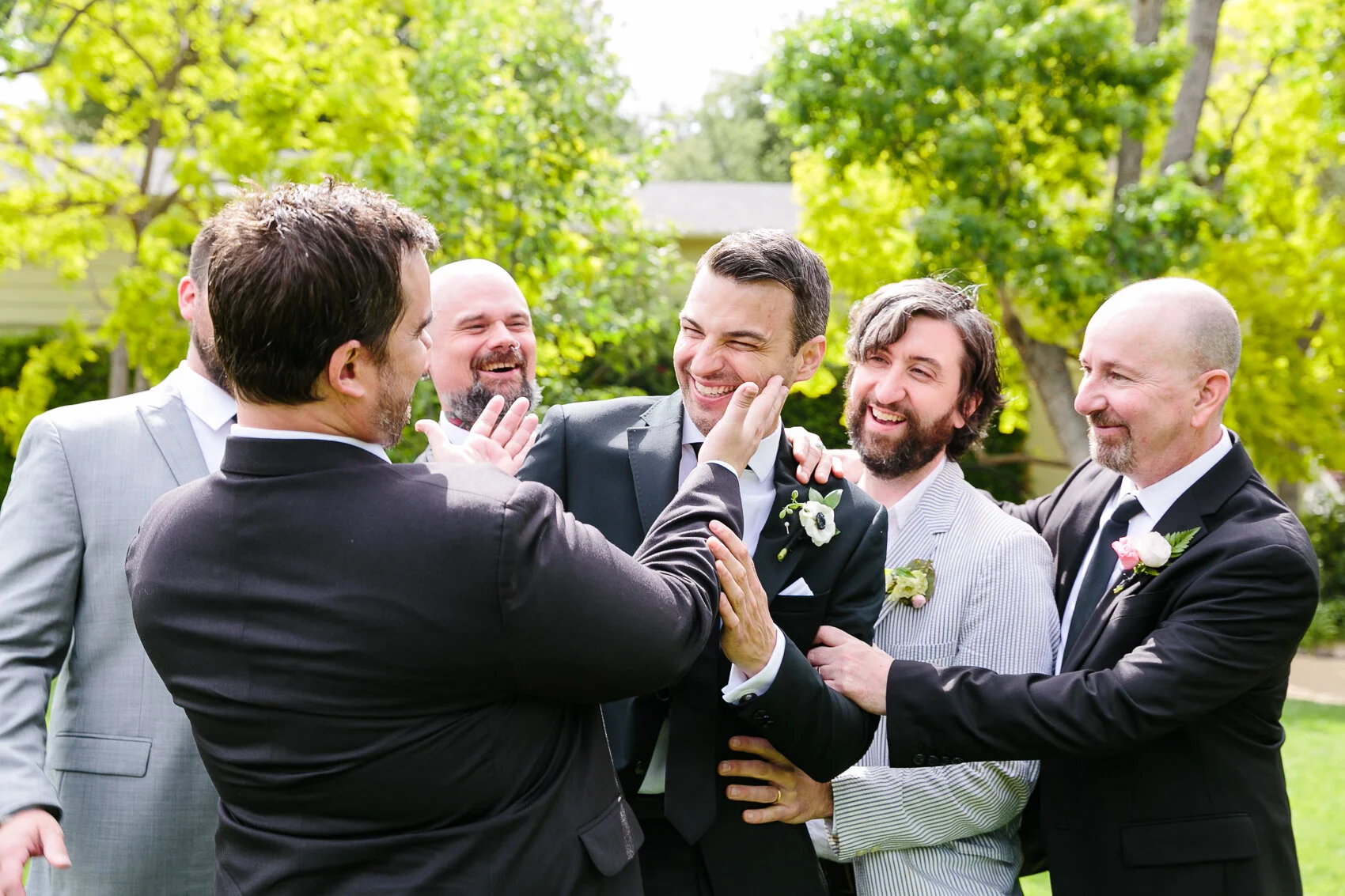 Group of men in suits laughing outdoors, wearing boutonnieres, in a celebratory setting.