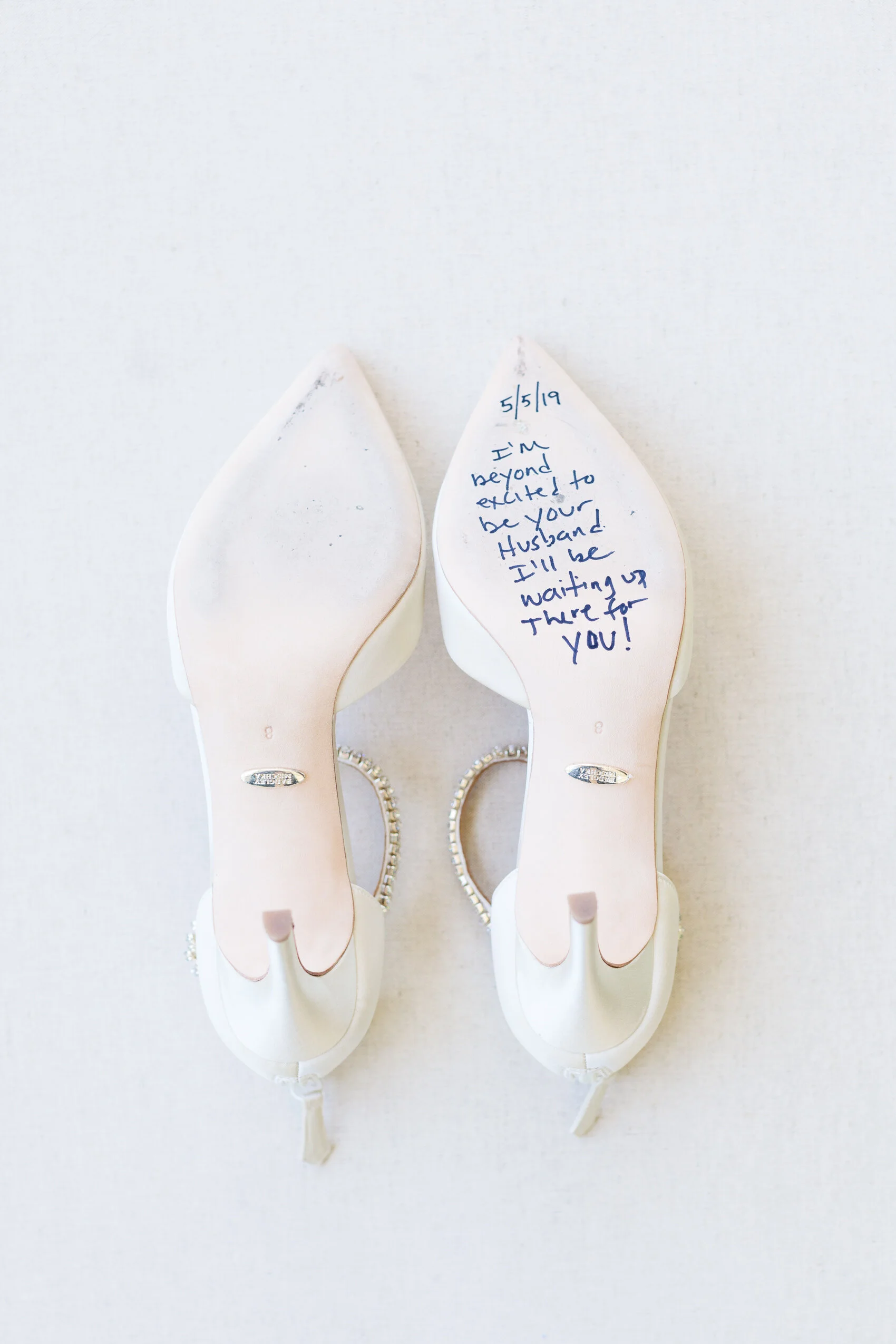 White bridal heels with a handwritten message on one sole saying, "5/5/19 I'm beyond excited to be your husband. I'll be waiting up there for you!"