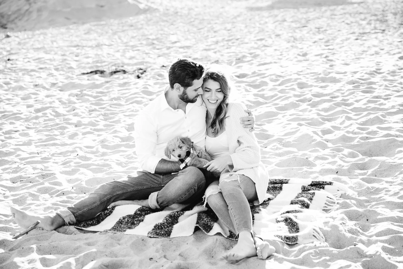 Couple sitting on a blanket at the beach with a small puppy, smiling in a black and white photograph.