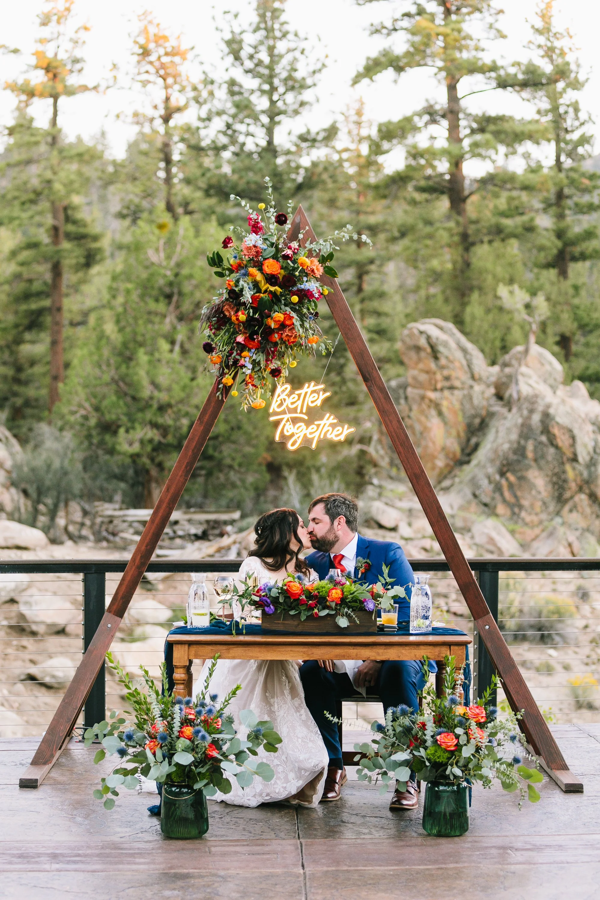 A couple kissing at a wedding reception table under a triangular wooden arch adorned with colorful flowers and a 'Better Together' neon sign, surrounded by forest scenery.