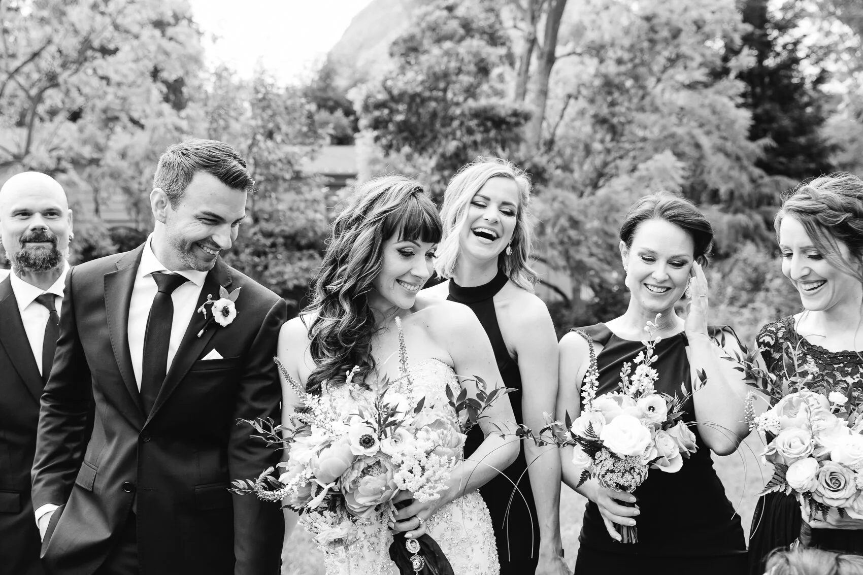 A black and white photo of a wedding party consisting of five people. They are dressed in formal attire, with the bride holding a bouquet and smiling. Other members include the groom and three bridesmaids, all laughing and smiling. They are outside w