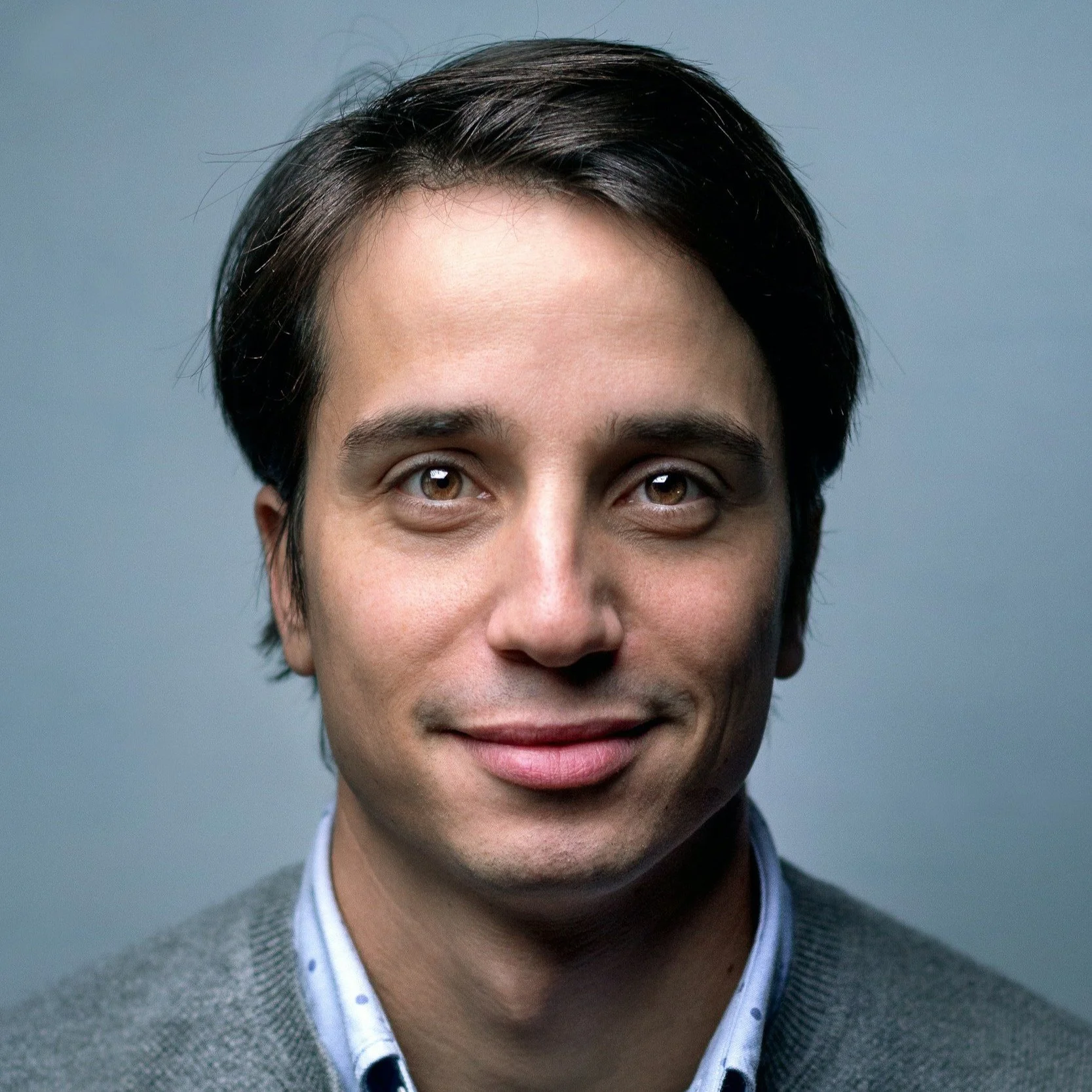 Close-up portrait of a young man with dark hair, light skin, and wearing a gray sweater over a collared shirt, smiling softly against a plain gray background.