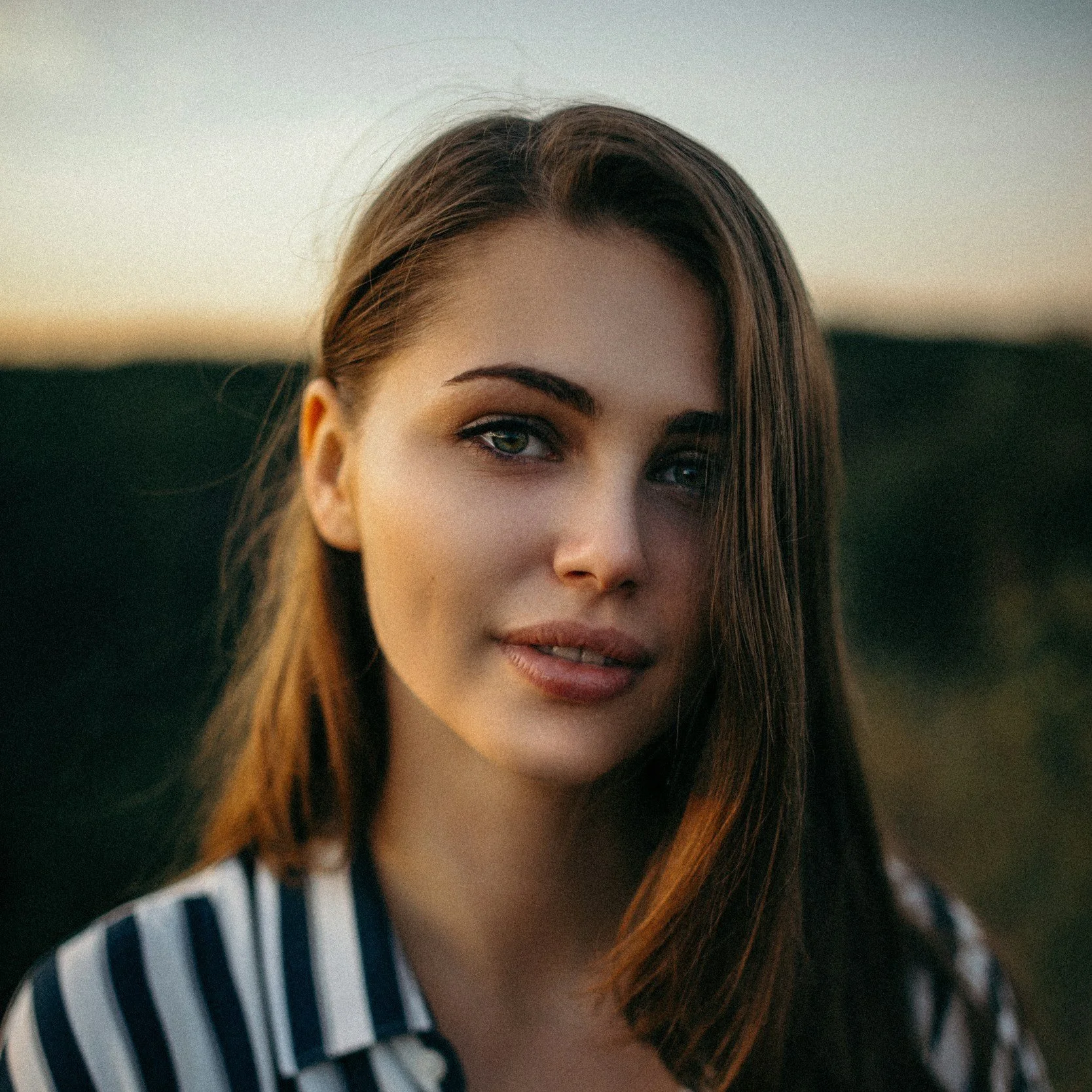 Close-up of a young woman with long brown hair and blue eyes, wearing a striped shirt, outdoors at sunset.