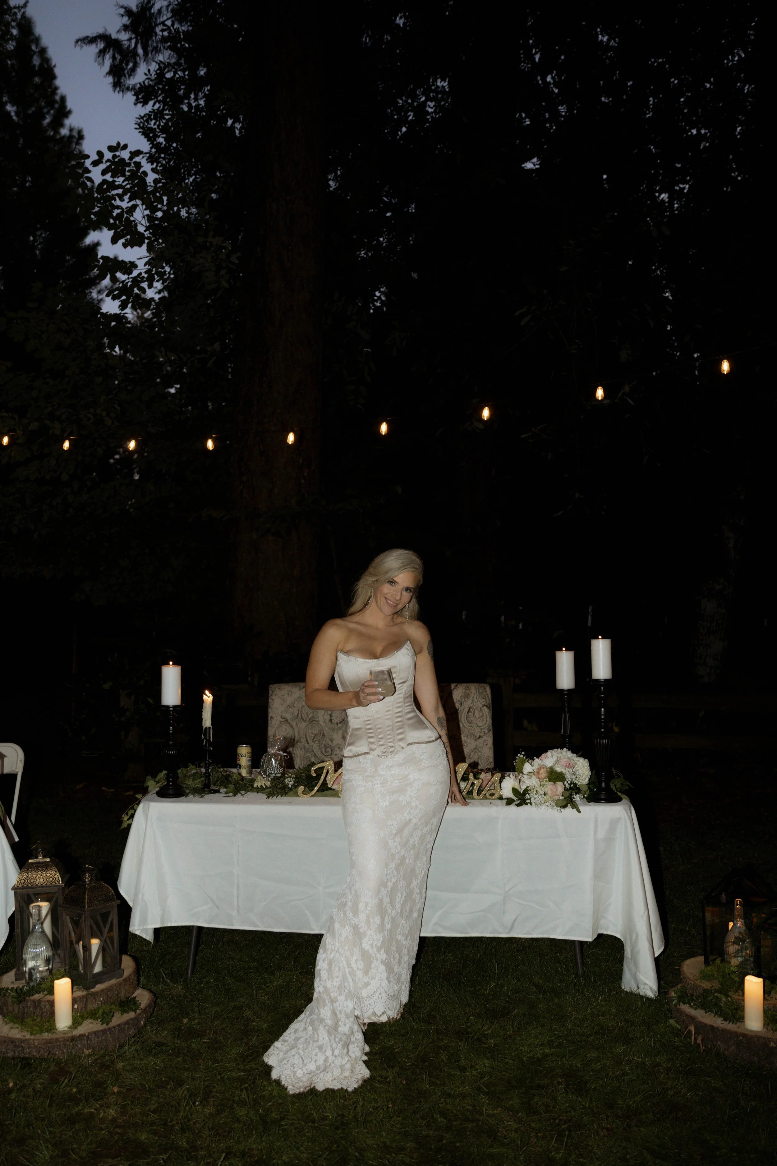 A bride standing at her sweetheart table underneath candles and string lights