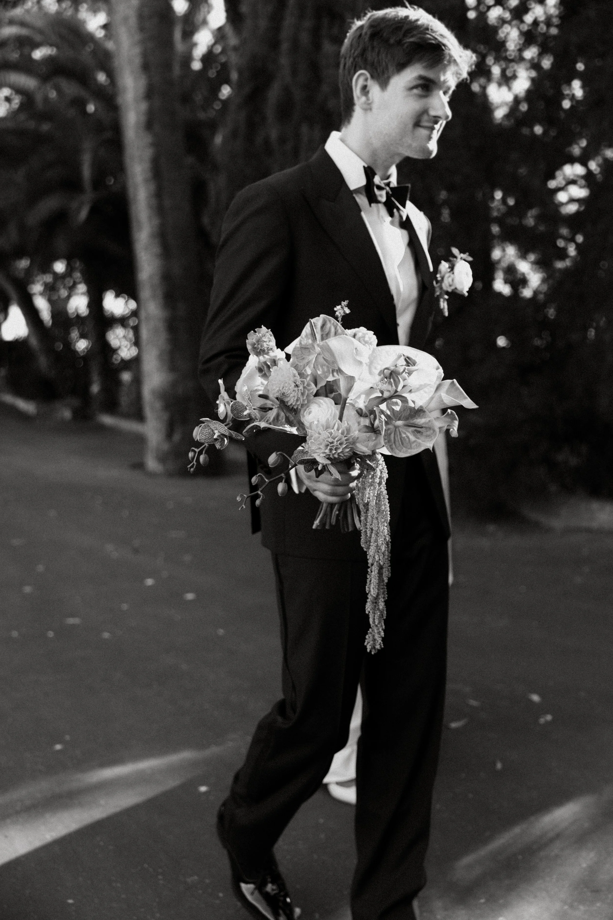 A young man dressed in a tuxedo holding a bouquet of flowers, standing outdoors at night with trees in the background.