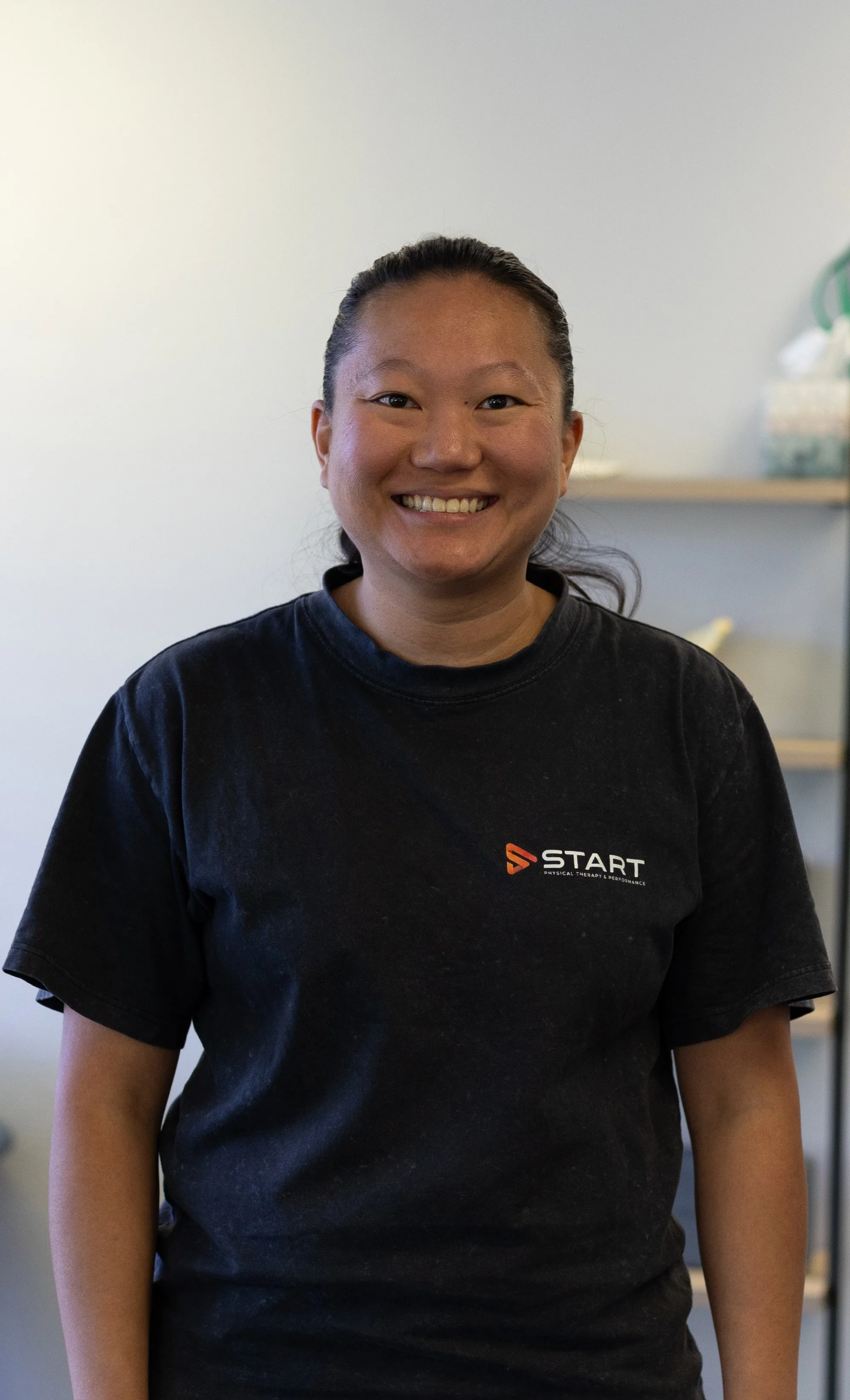 Smiling woman with dark hair tied back, wearing a black T-shirt with the 'START' logo, standing indoors with shelves and supplies in the background.