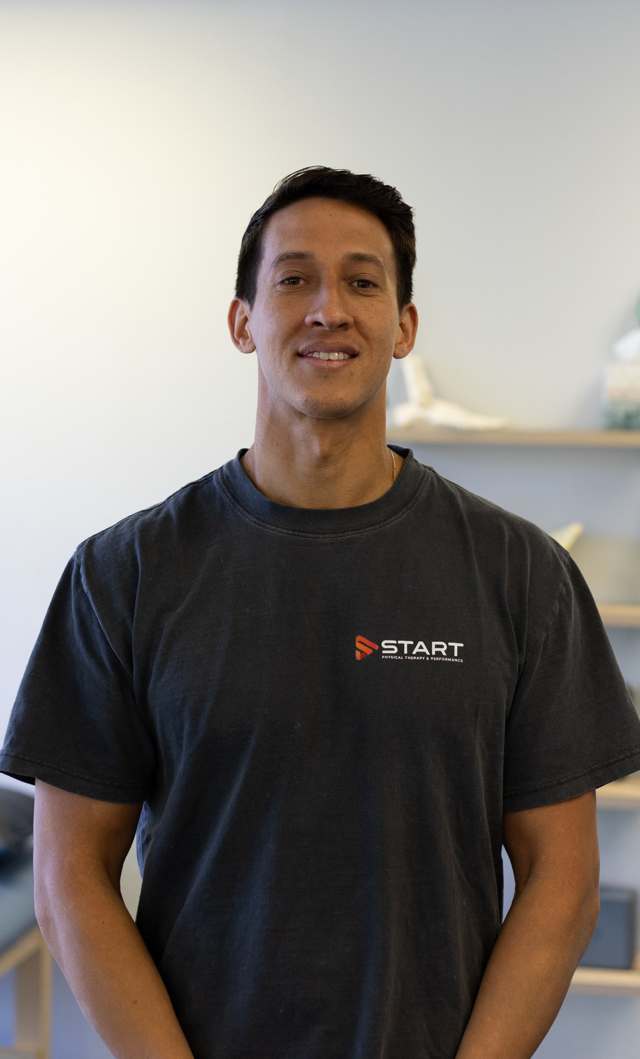 A young man with short dark hair smiling, wearing a black T-shirt with a 'START' logo, standing indoors against a plain background with some shelves and objects.