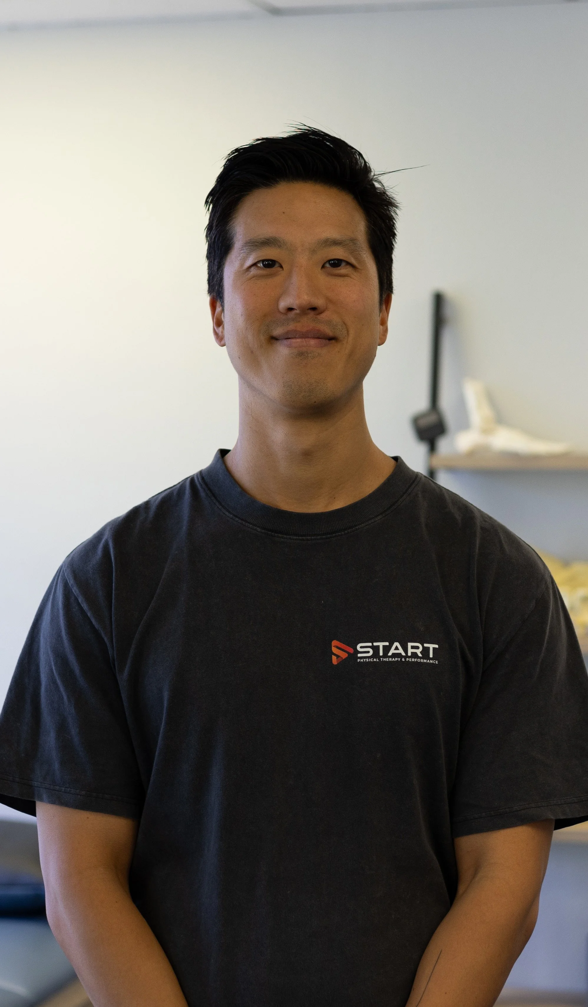 A young man with short black hair and a slight smile, wearing a black T-shirt with 'START' logo, standing indoors in front of a neutral background.