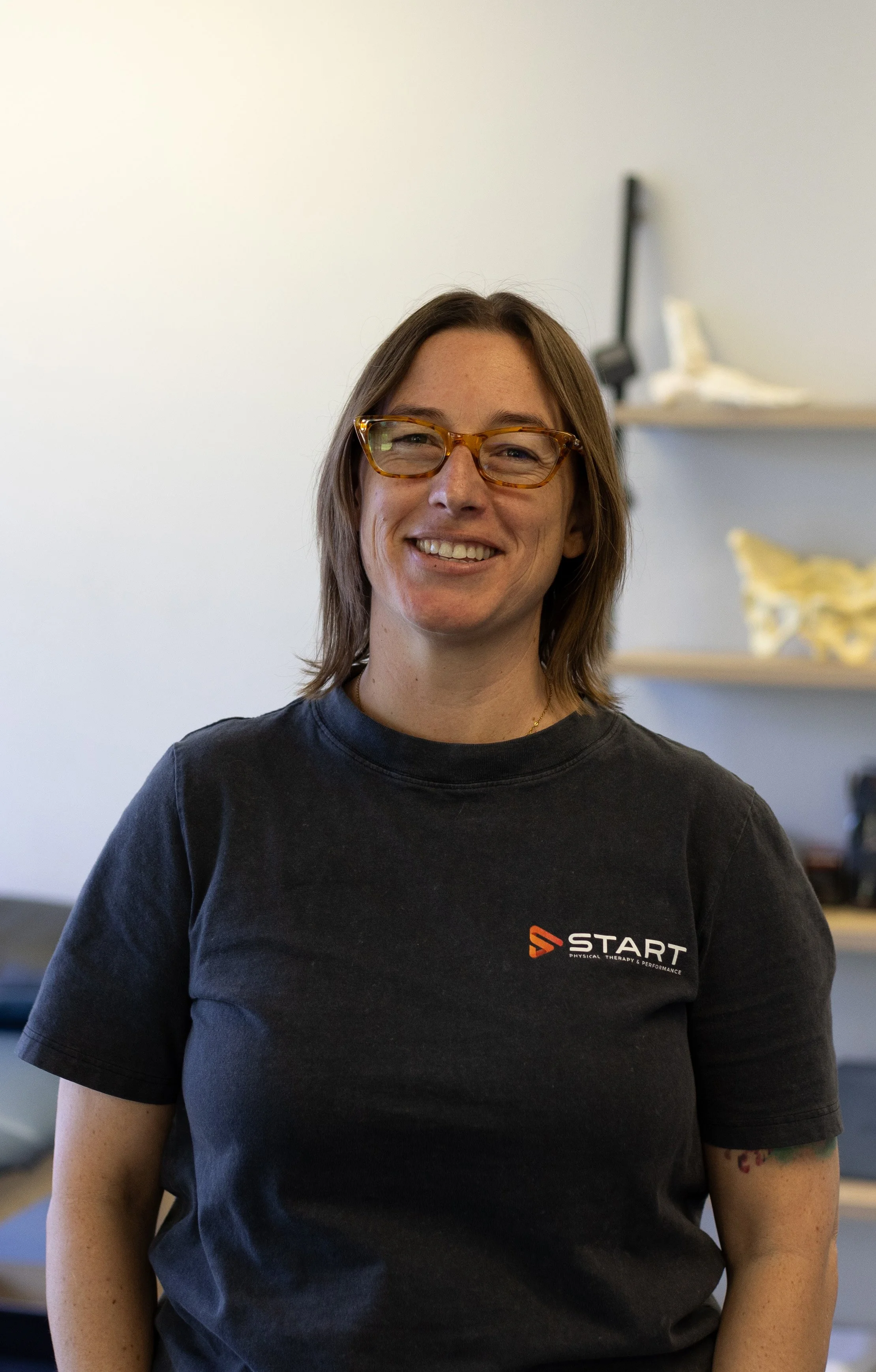 A woman with shoulder-length brown hair, wearing glasses and a black T-shirt with a logo on the front, smiling in an indoor setting with a white wall and shelves behind her.