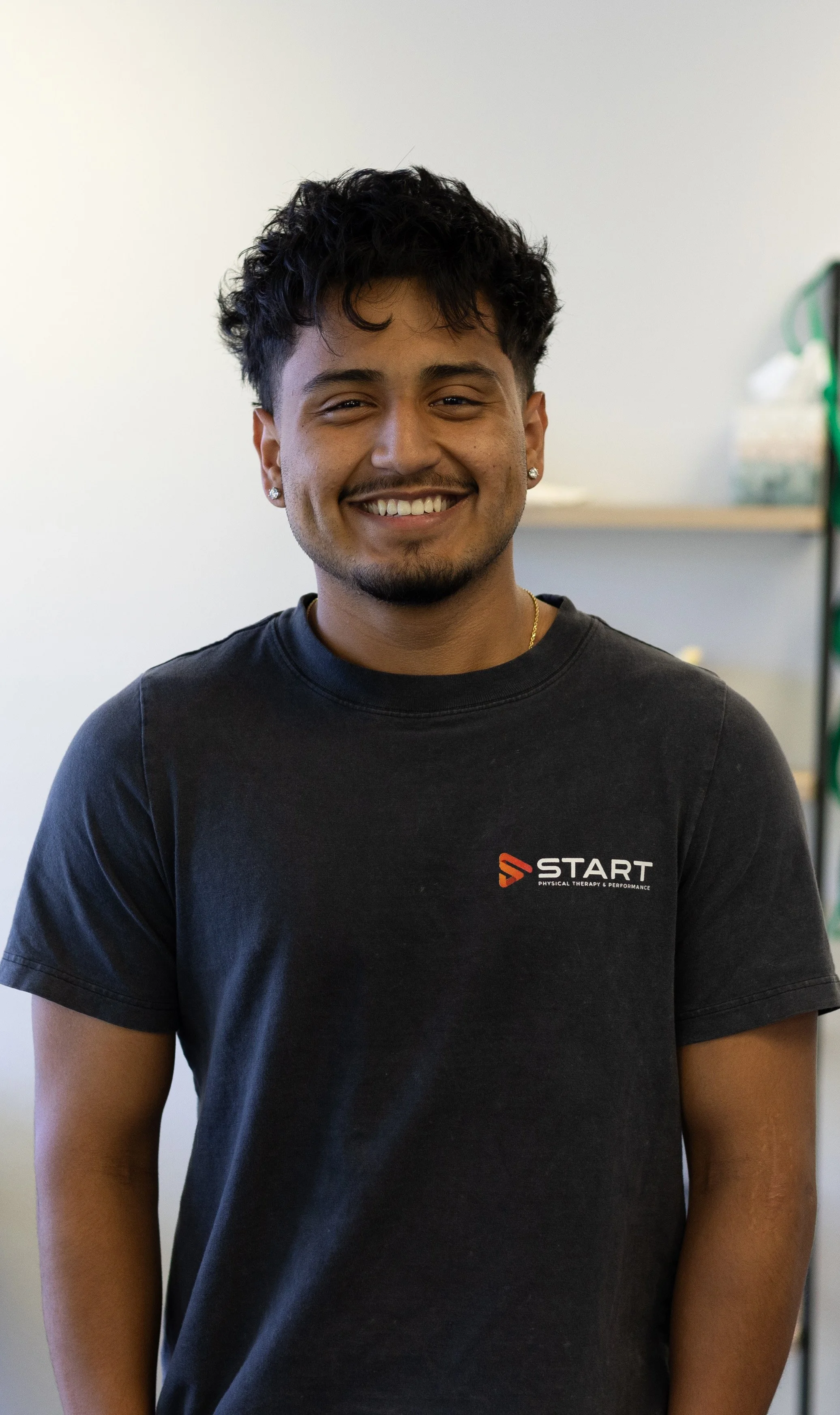 A smiling young man with dark curly hair, wearing a black T-shirt with a logo that reads 'START Physical Therapy & Performance,' standing inside with a light-colored wall in the background.