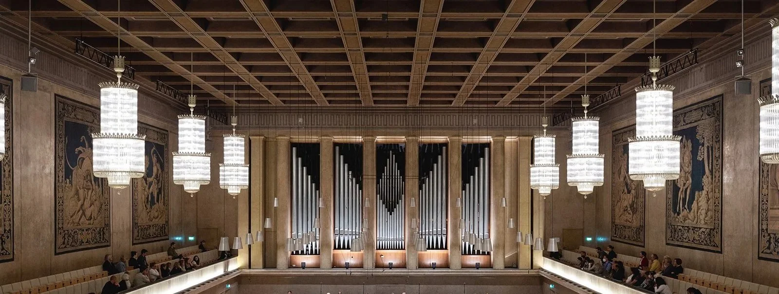 Interior of a concert hall with large hanging chandeliers, an organ with many pipes at the front, ornate tapestries on the walls, and audience seating along the sides.